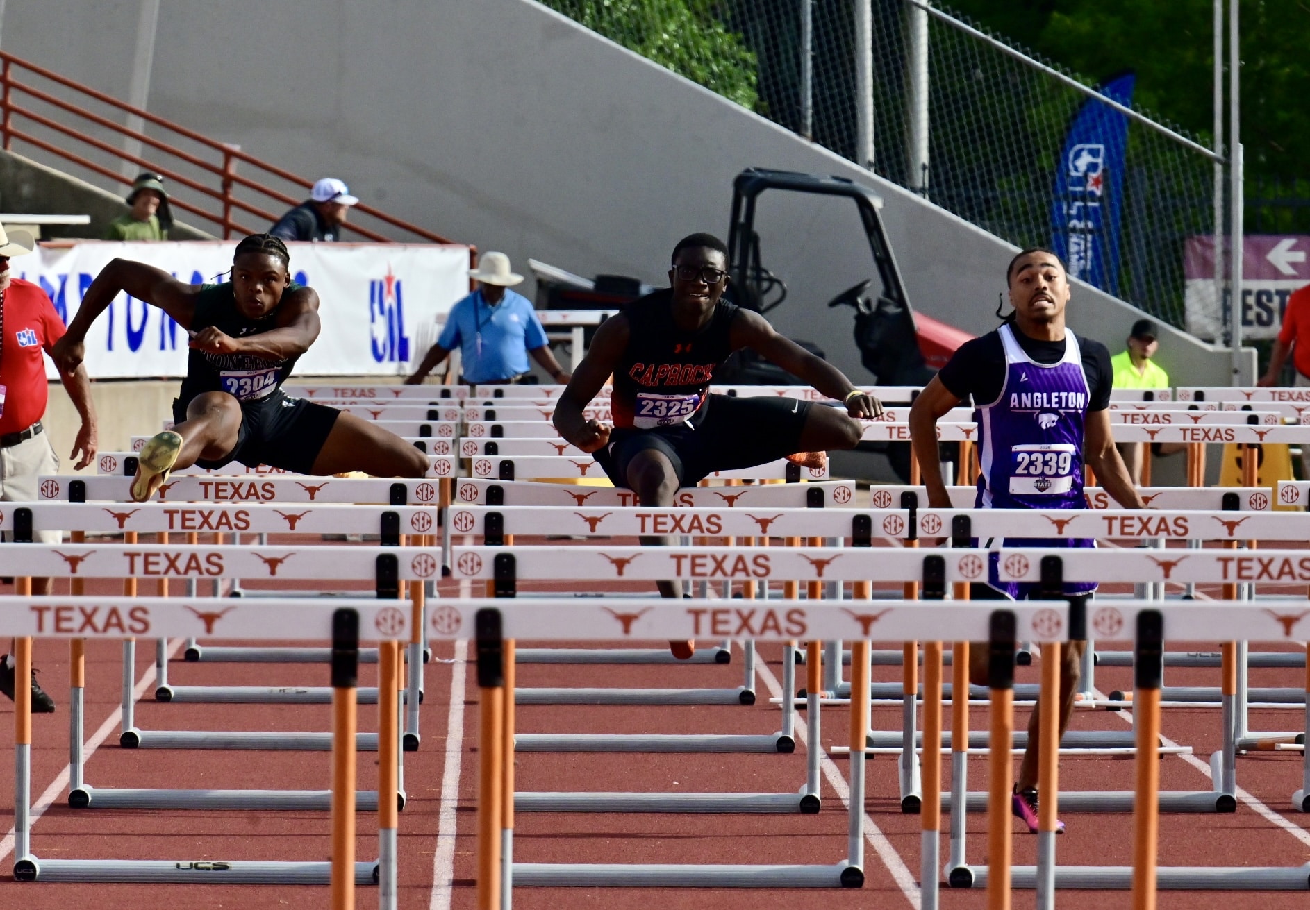Three male hurdlers jump over Texas hurdles on a track, mid-race action with banners and officials in the background