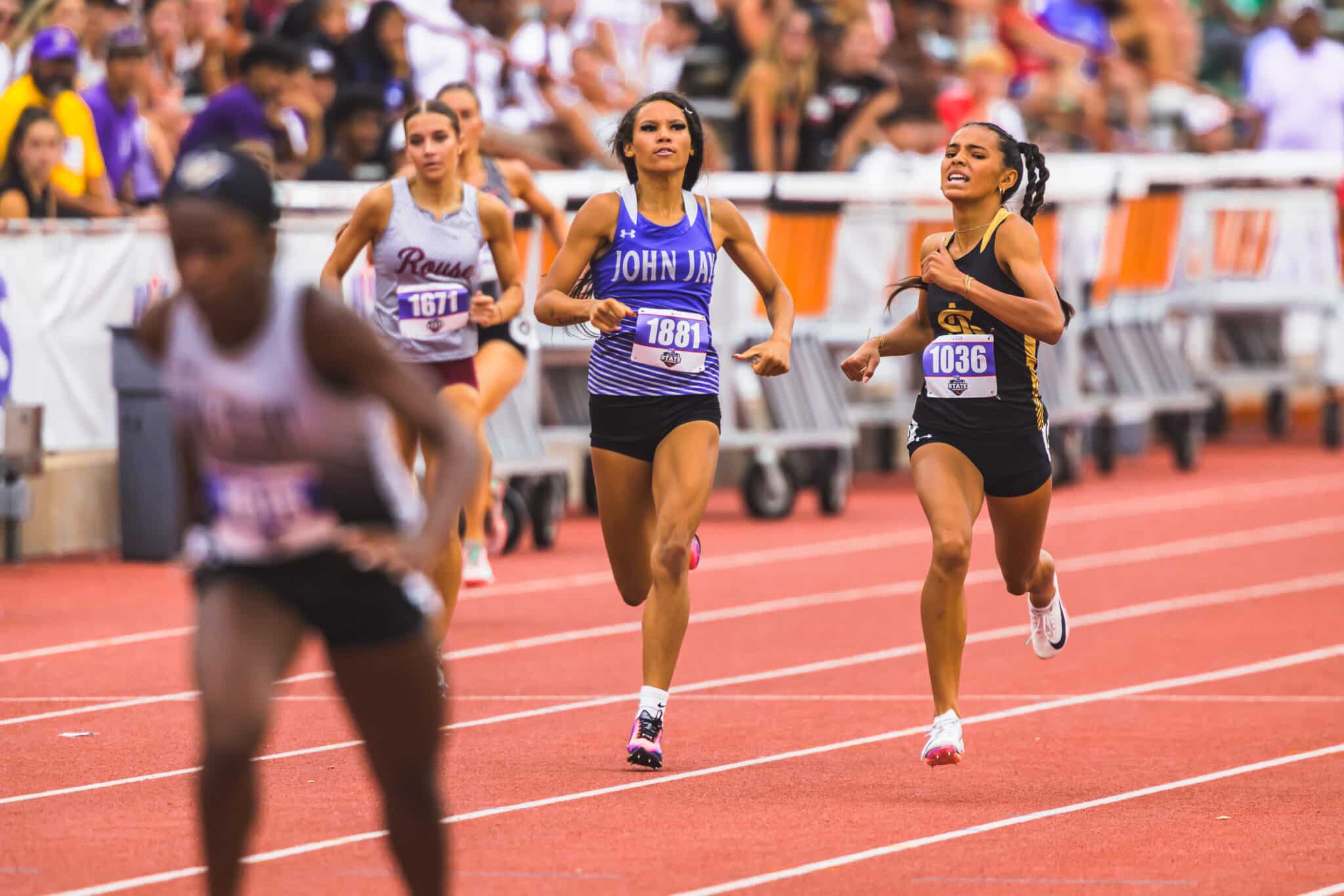 Female sprinters in a crowded stadium race on a red track; central runner in blue (John Jay) with bib 1881, right runner in black with bib 1036.