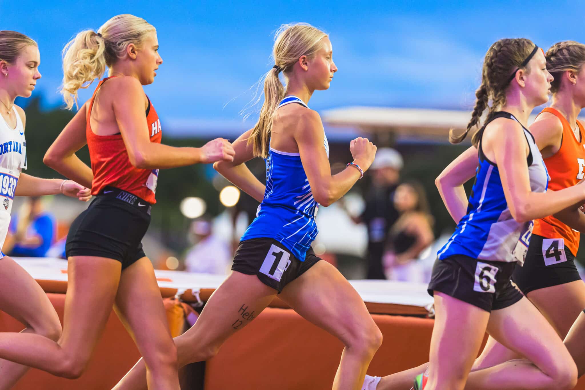 Group of female runners sprinting on an outdoor track, wearing numbered bibs and athletic gear, mid-race.