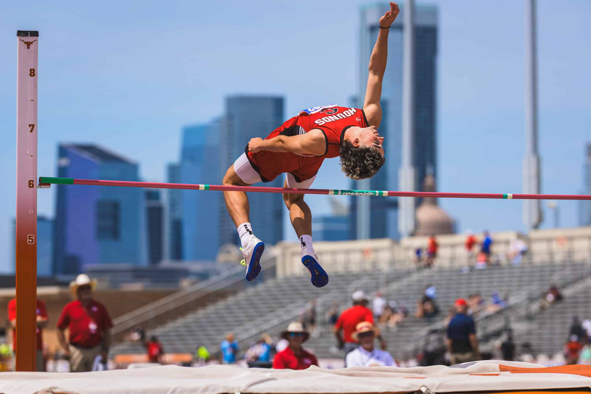 Male high jumper in a red uniform clears a bar during a track and field event in a city stadium with skyscrapers in the background.