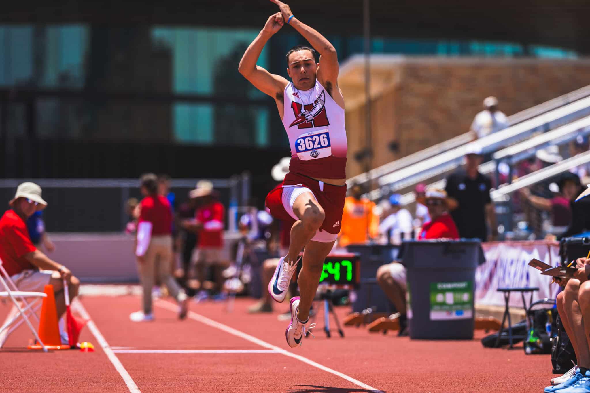 Male hurdler mid-jump over track hurdles during a race, bib 3626, in a crowded stadium.