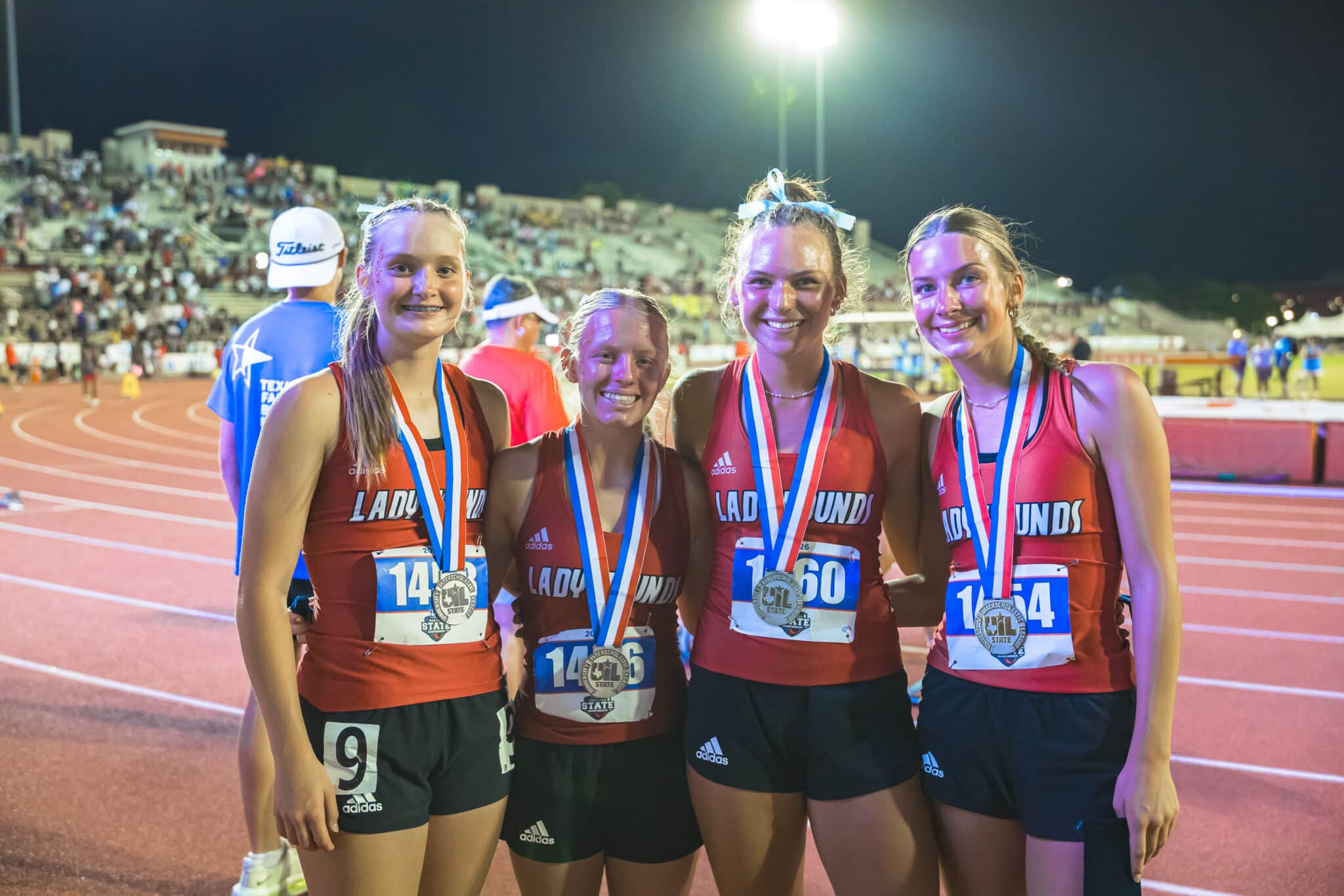 Four young female runners in red uniforms pose with medals on a track at night, stadium lights behind them.