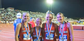 Four young female runners in red uniforms pose with medals on a track at night, stadium lights behind them.