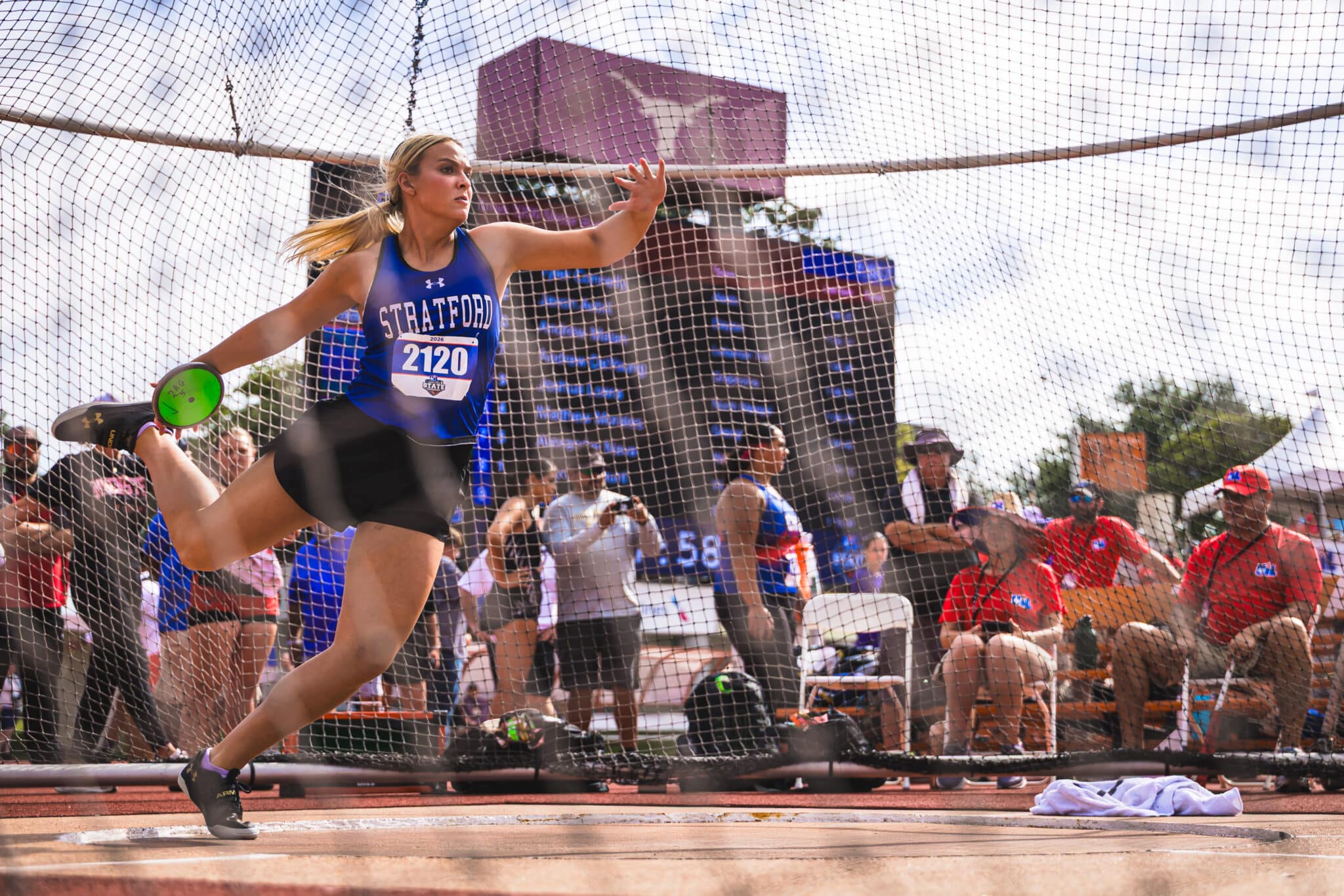 Female discus thrower in a blue Stratford uniform mid-throw, holding a green discus inside the safety netting at a track meet.