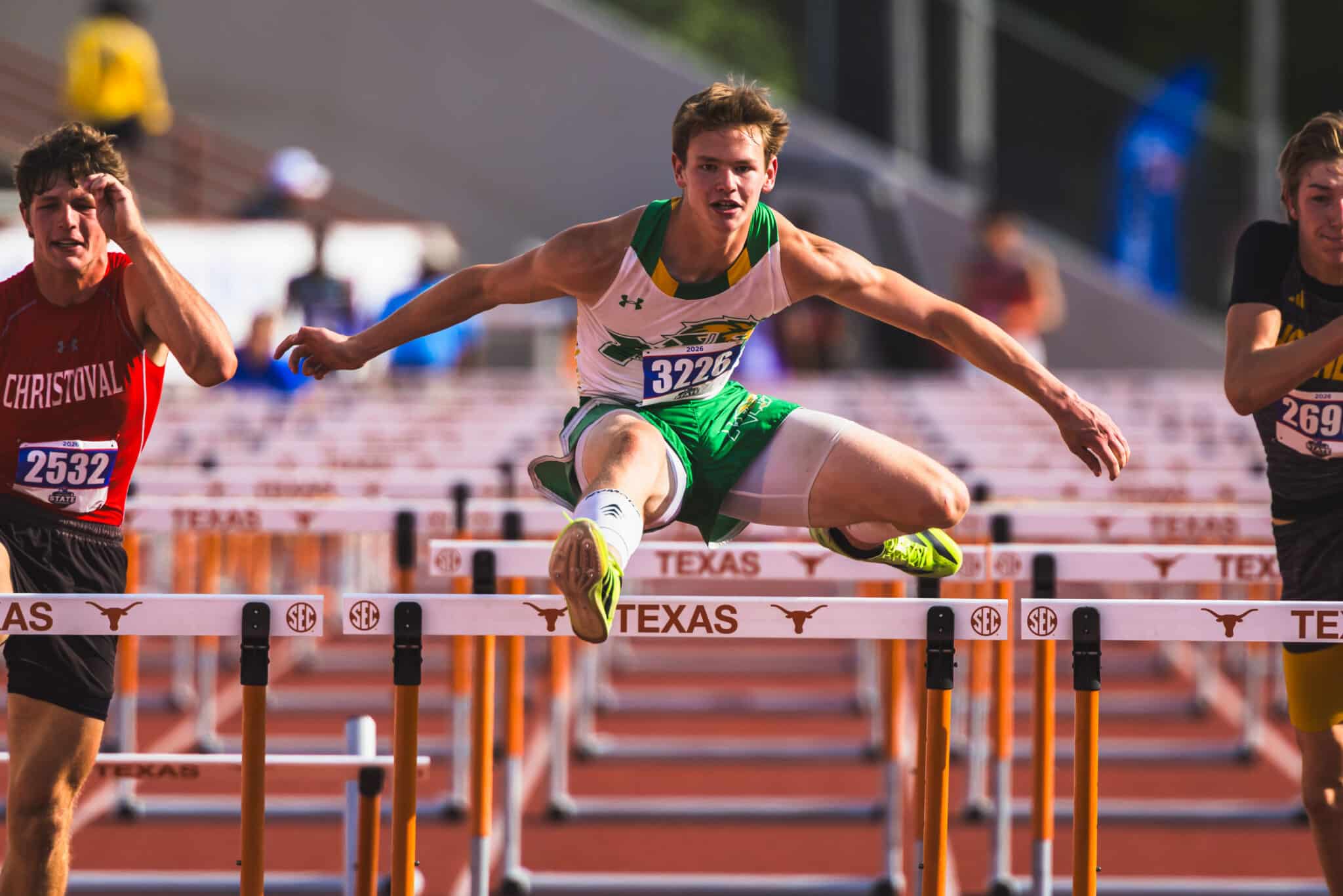 Male hurdler in green-and-white uniform clears a Texas SEC hurdle mid-air during a race on the track
