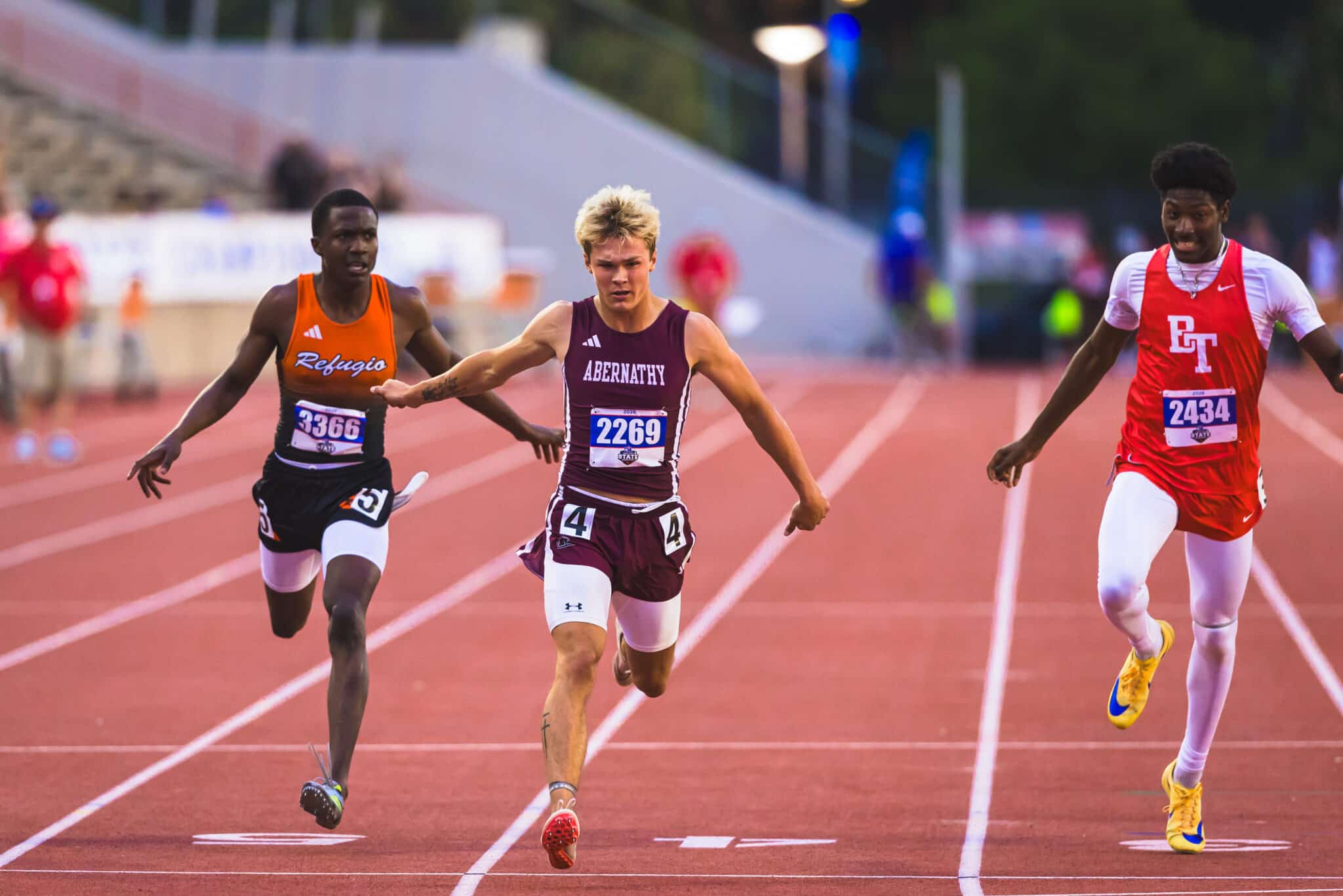 Three male sprinters race down a track, center athlete in maroon Abernathy uniform (bib 2269) ahead, flanked by a left runner in orange Refugio (bib 3366) and a right runner in red CT (bib 2434).