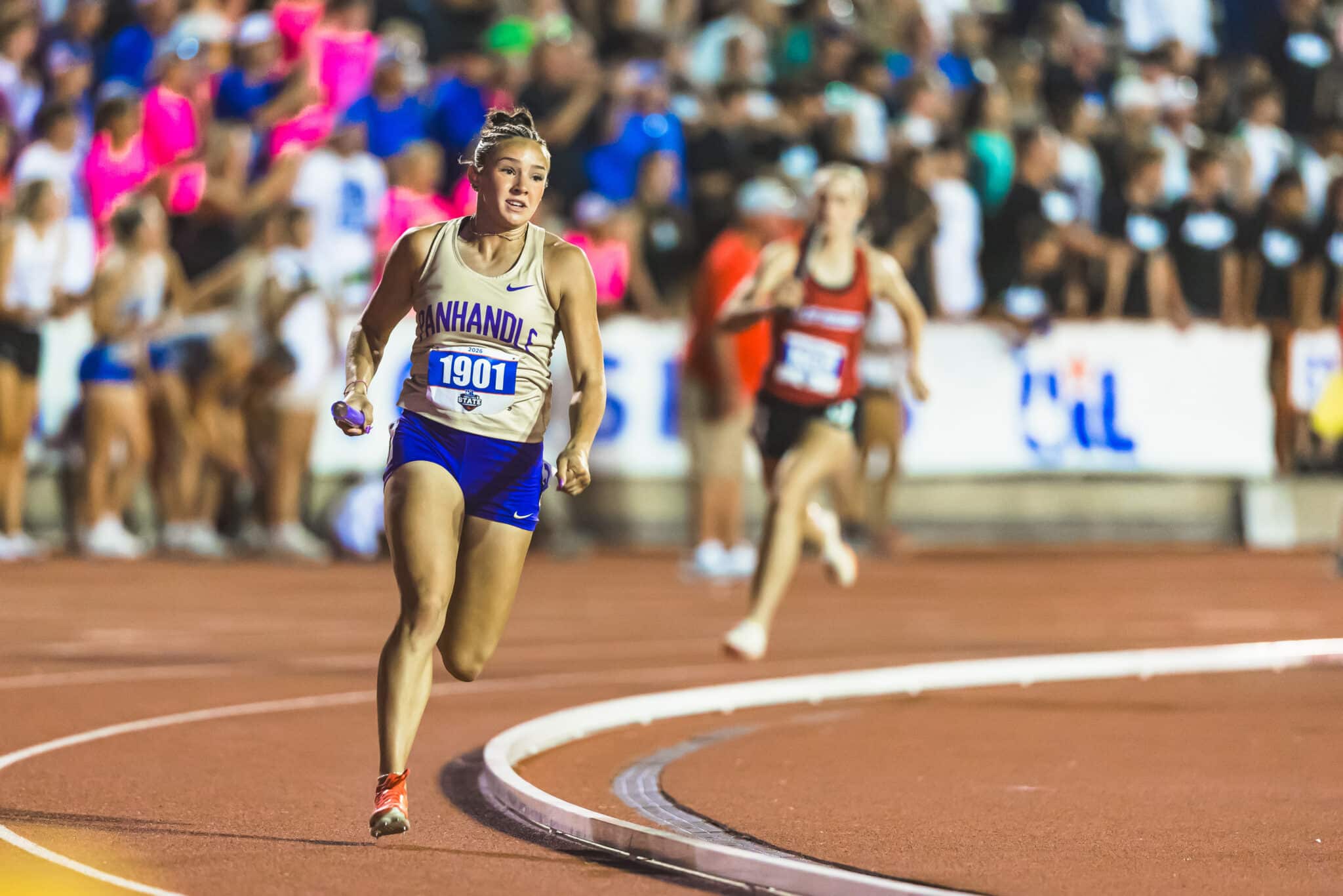 Female sprinter in beige top and blue shorts runs on a curved red track, bib 1901, with a cheering crowd in the stands behind.