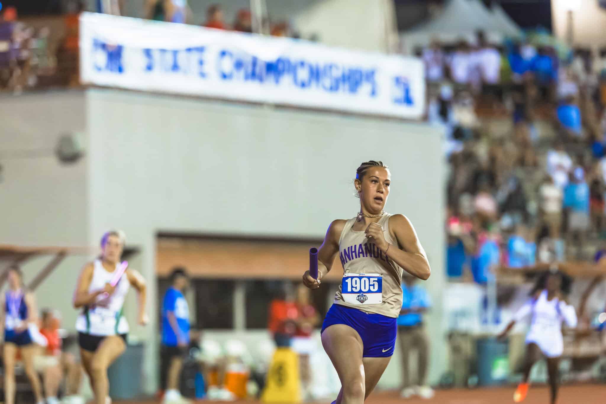 Female sprinter in beige top and blue shorts running with a purple baton during an indoor track meet, with a cheering crowd and banner in the background (bib number 1905).