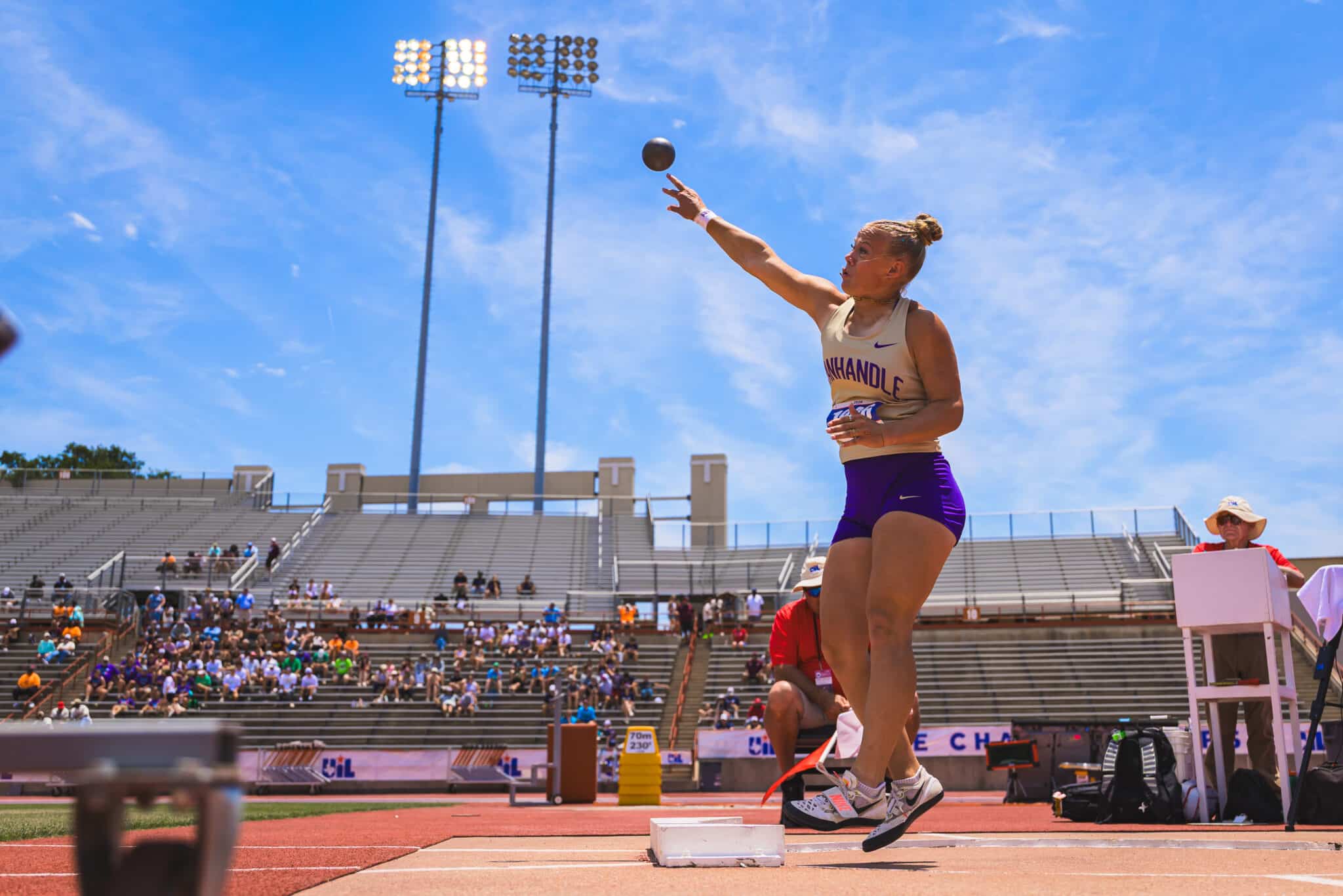 Female track and field athlete mid-jump on a long jump runway, sky-blue background and stadium stands full of spectators behind her.