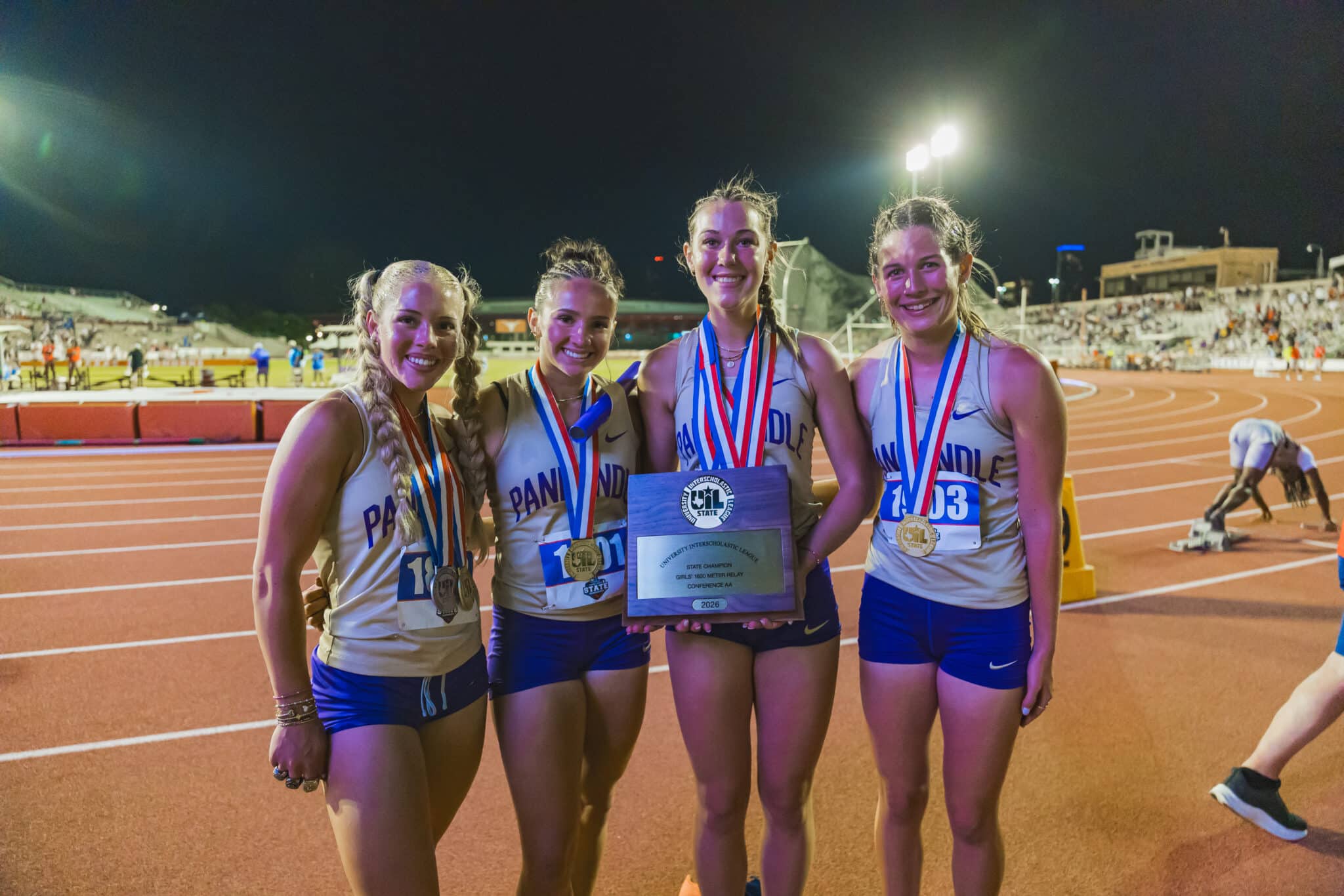 Four smiling female runners in matching uniforms pose with medals and a trophy on a nighttime outdoor track field.