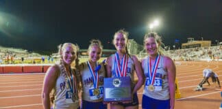 Four smiling female runners in matching uniforms pose with medals and a trophy on a nighttime outdoor track field.