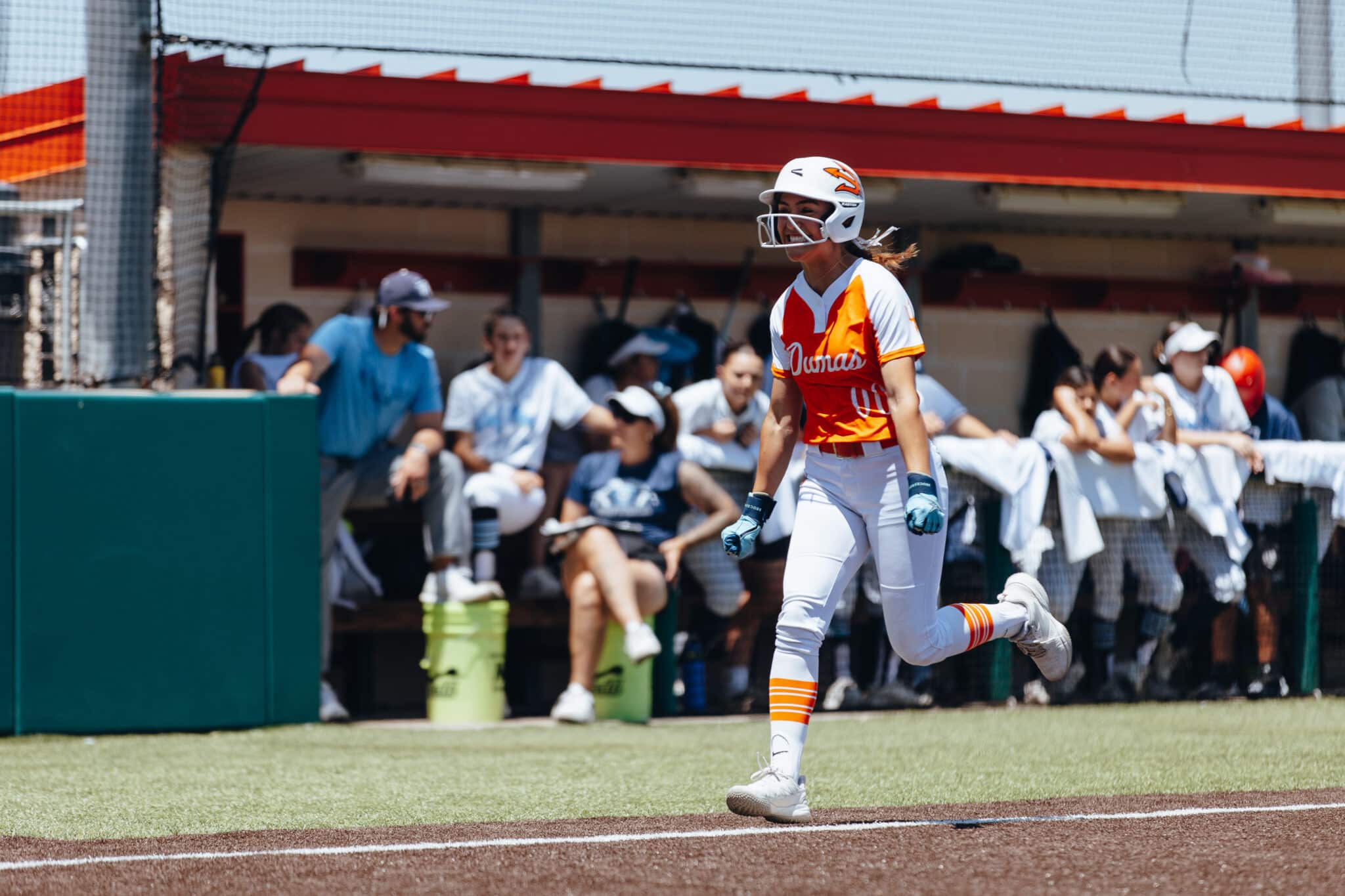 Female softball player in orange-and-white uniform runs on the field, wearing a helmet, with fans watching from the dugout behind her.