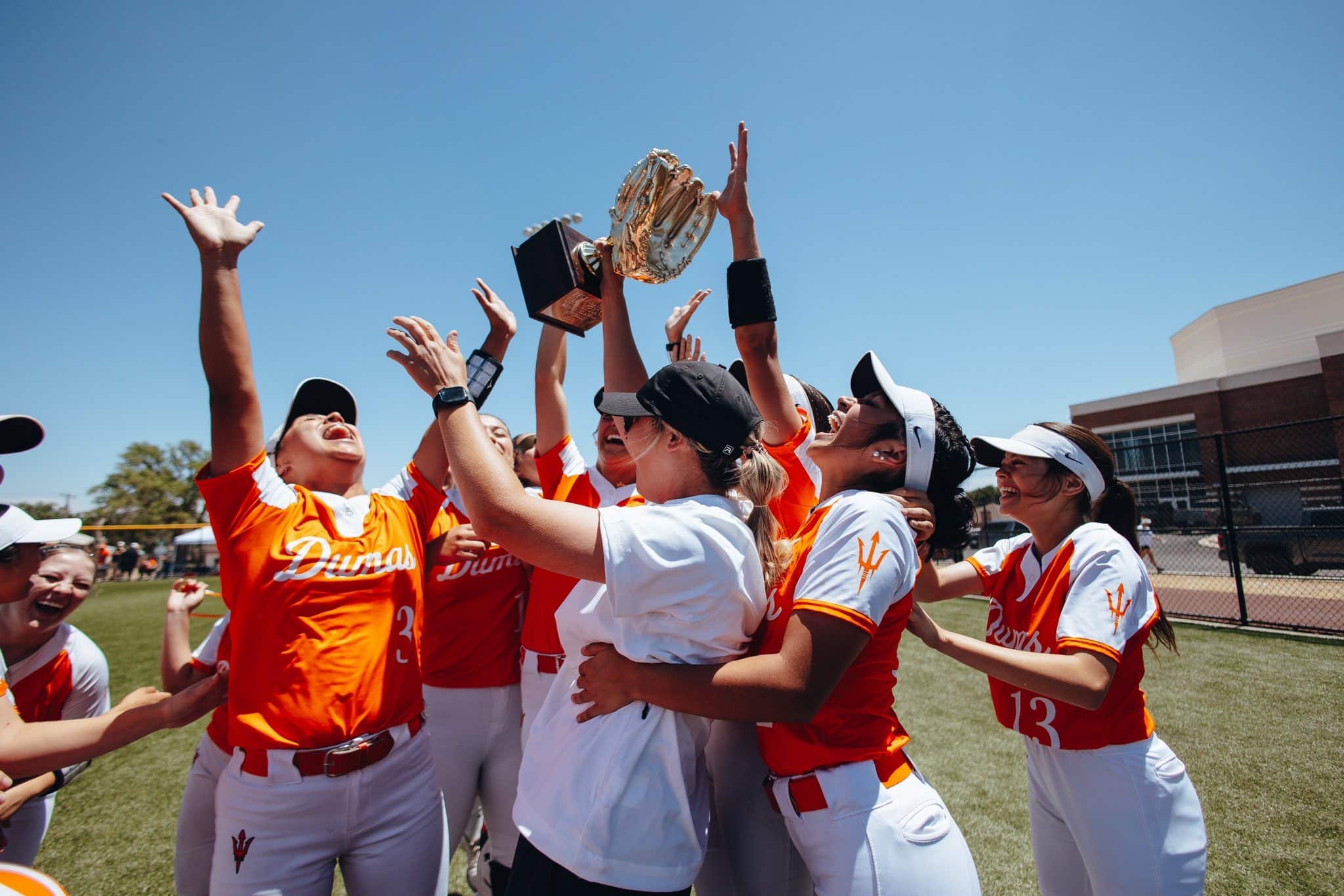 Girls' sports team in orange uniforms lifts a trophy in celebration on a sunny day at the field.