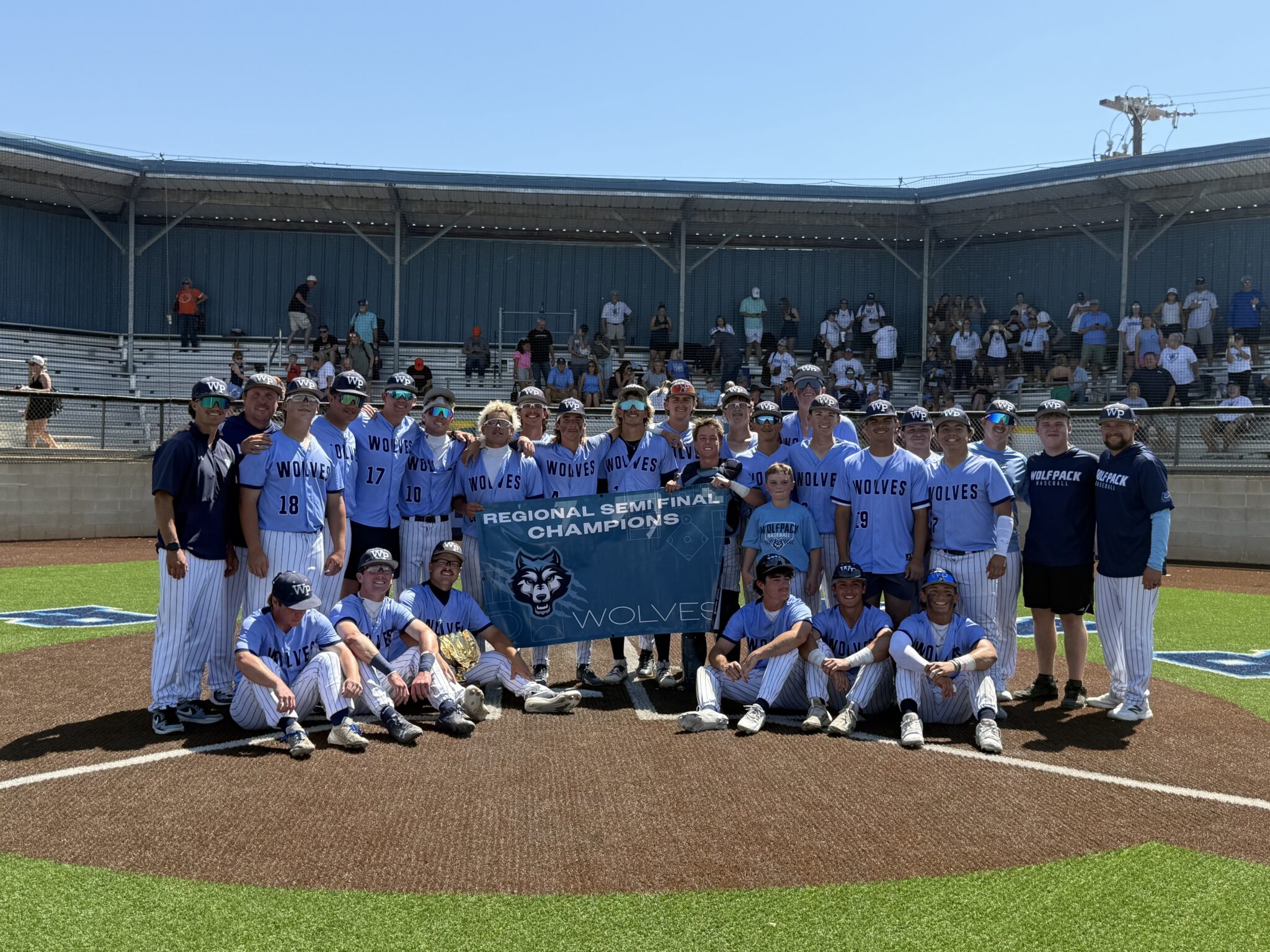 Group of baseball players in light blue uniforms posing on the field with a banner that reads'Regional Semifinal Champions'. Behind them are spectators in bleachers under a clear blue sky.