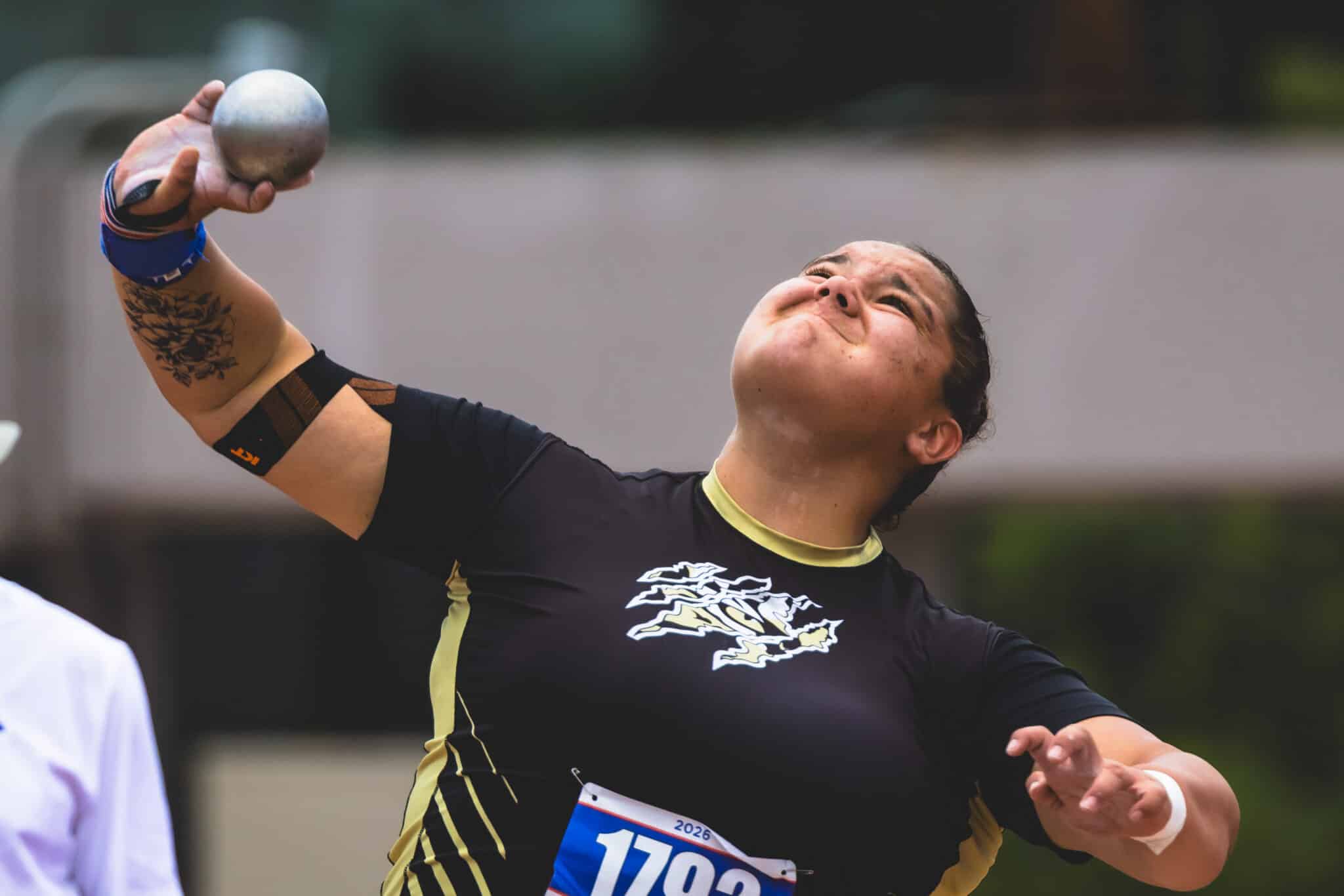 Female athlete in black and gold jersey releasing a shot put, tattoo on left forearm visible.