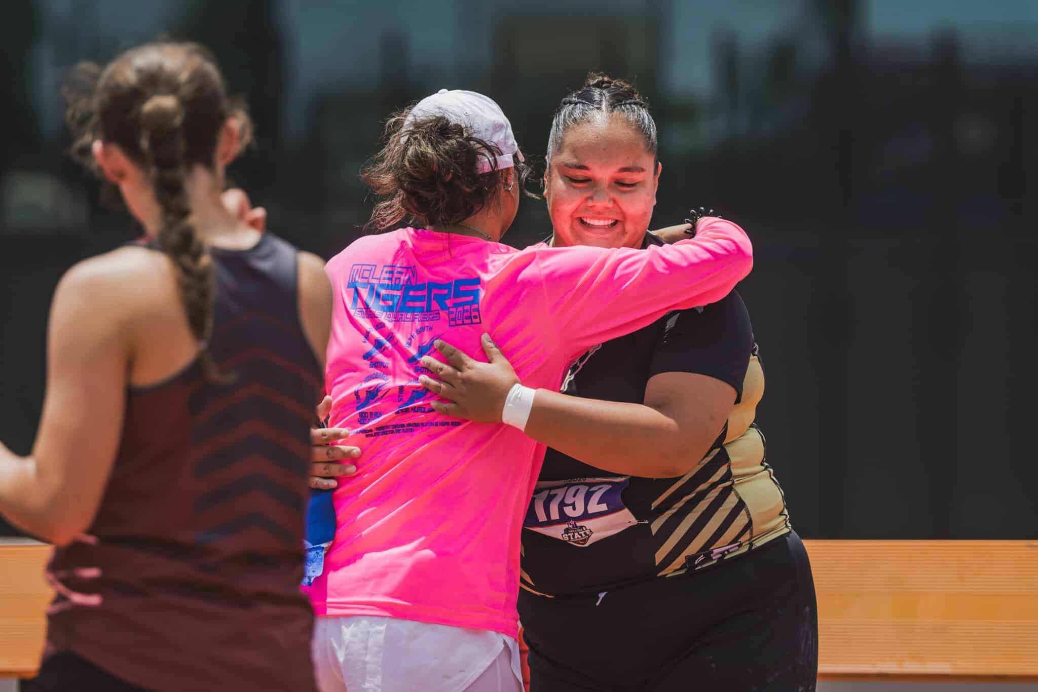 Two female runners share a warm hug after a race; one wears a bright pink shirt with a printed graphic and a bib number visible (792).