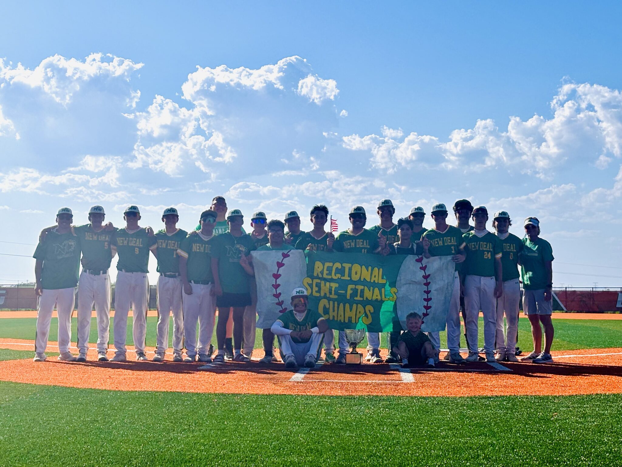 Group of baseball players in teal uniforms pose with banners and a trophy on a baseball field under a blue, cloudy sky.
