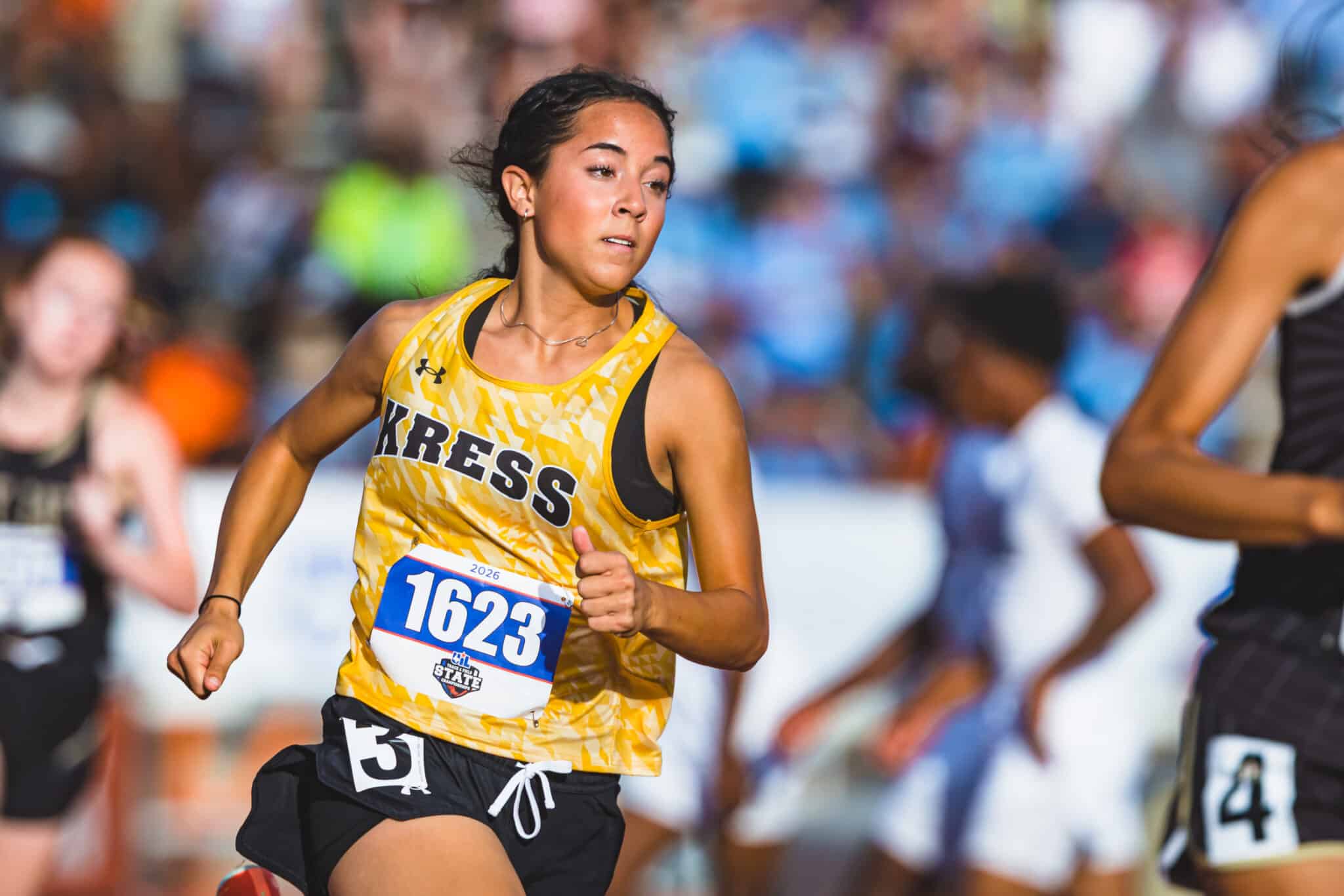 100A2513 | Press Pass Sports Female runner in a yellow patterned sleeveless top, bib 1623, mid-race with a crowd in the blurred background.