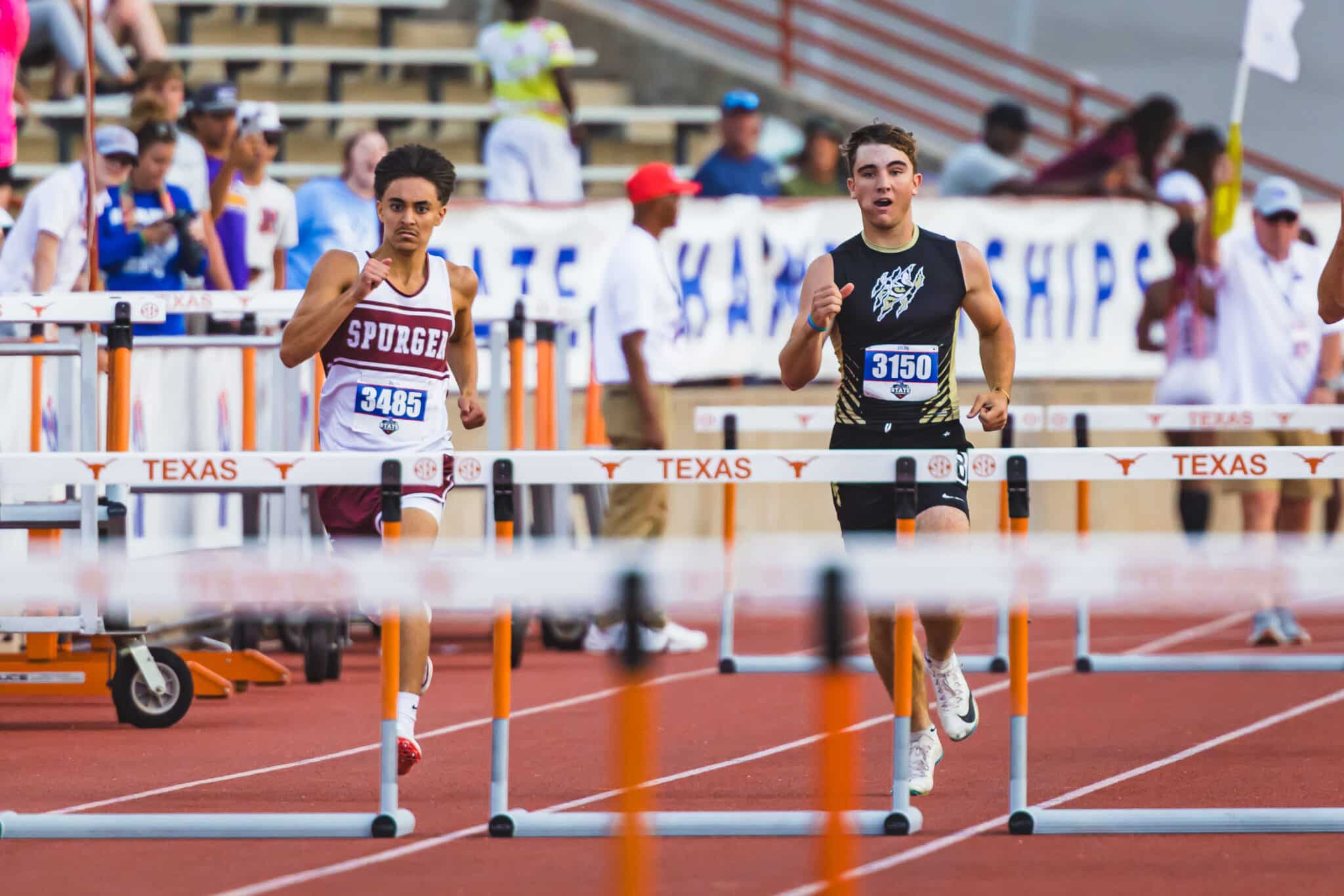 100A3039 | Press Pass Sports Two male hurdlers sprint toward the finish line on a red track, mid-race, with hurdles and a Texas banner in the background.