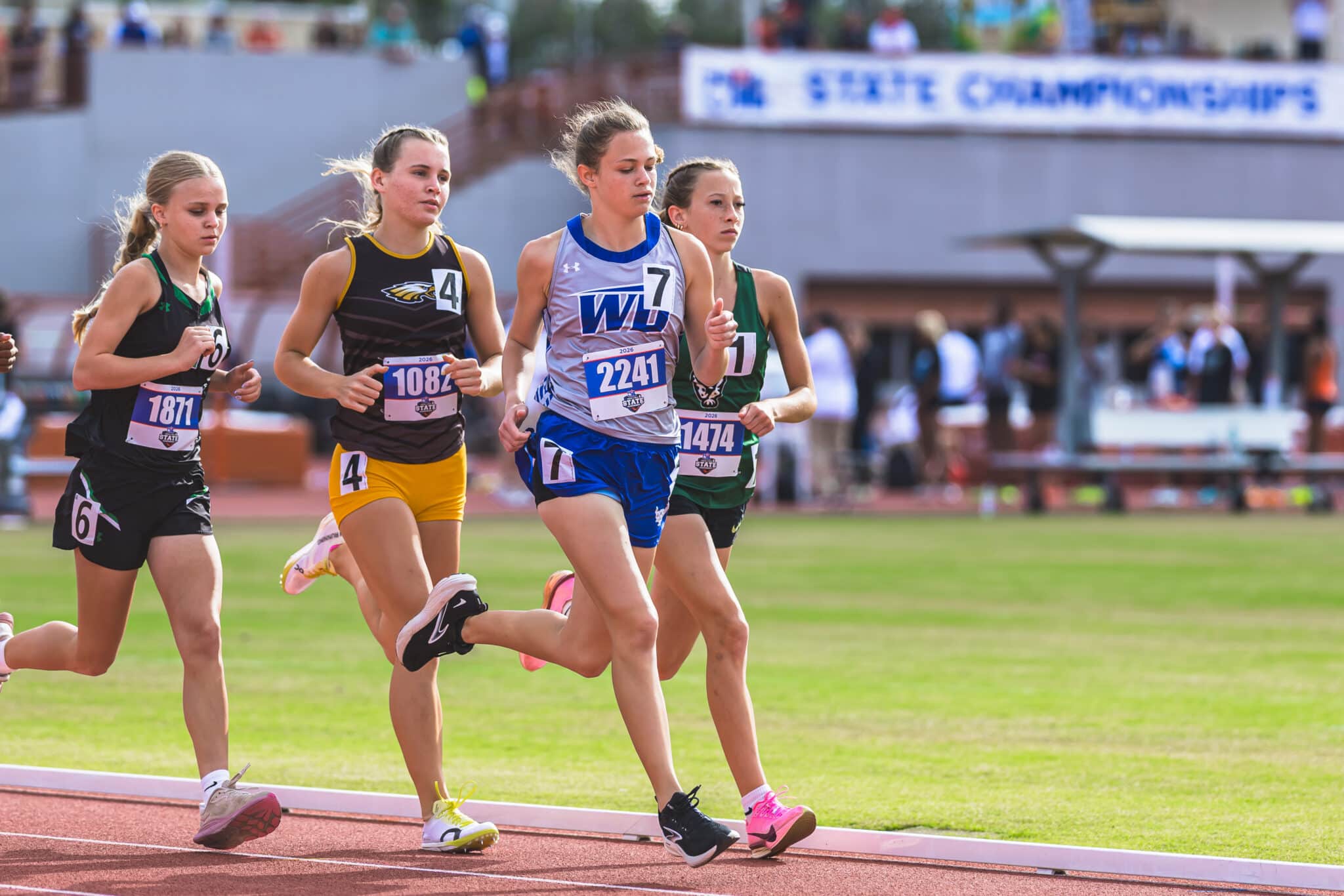 100A7409 | Press Pass Sports Group of female sprinters running on a track, bib numbers visible, green field in the background.
