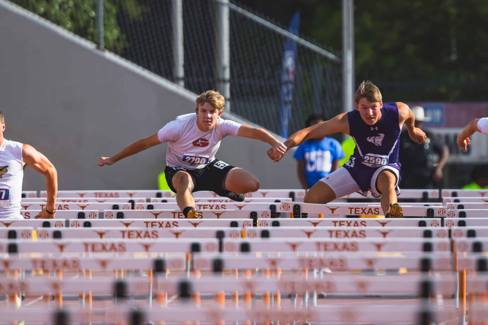 100A1300 | Press Pass Sports Runners in a hurdles race leaping over orange-and-white Texas hurdles on an outdoor track, focused expressions and athletic motion as they clear the barrier beside each other.