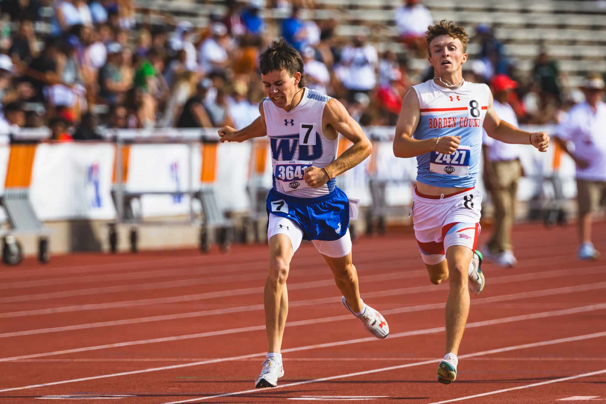 100A0816 | Press Pass Sports Two male sprinters mid-race on a red track, bib numbers 7 and 8, with blurred spectators behind them in the stands.