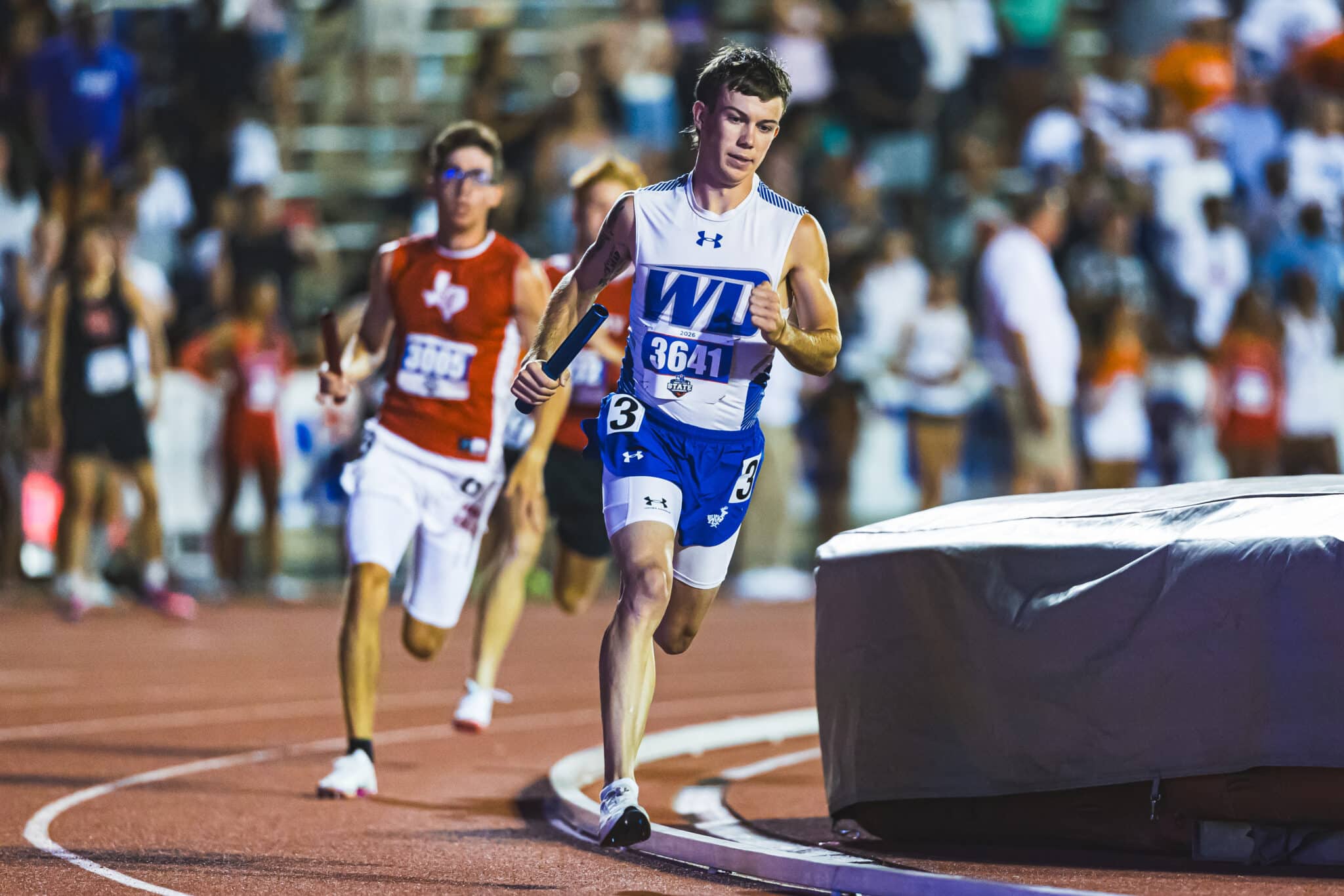 100A4323 | Press Pass Sports Male sprinter in white and blue uniform with bib 3641 runs on a track, holding a baton as crowd watches in the blurred background.