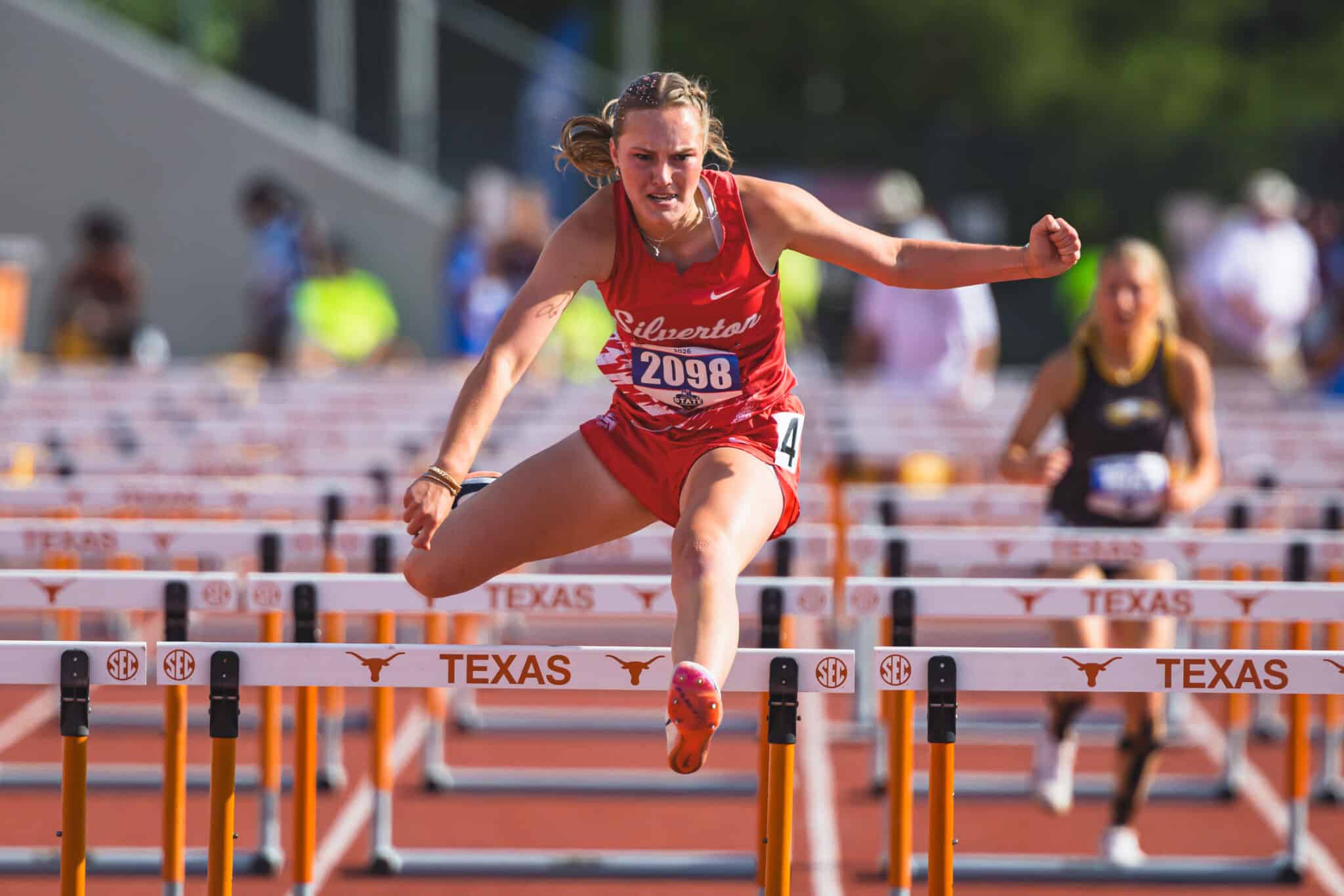 100A1207 | Press Pass Sports Female sprinter in red uniform clearing a hurdle during a Texas track race, intense concentration on her face.