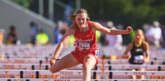 Female sprinter in red uniform clearing a hurdle during a Texas track race, intense concentration on her face.
