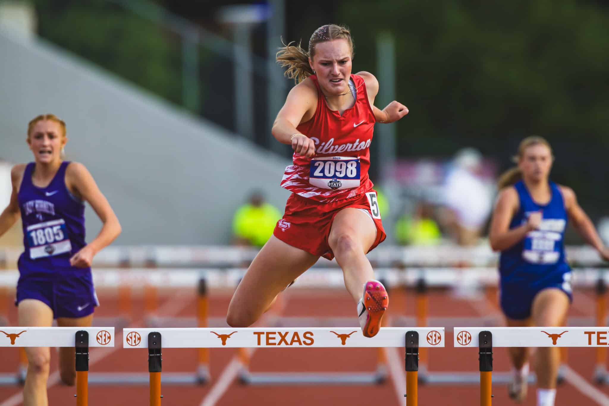 100A2849 | Press Pass Sports Female hurdler in red clears a hurdle during a track race, Texas banner visible in the foreground.