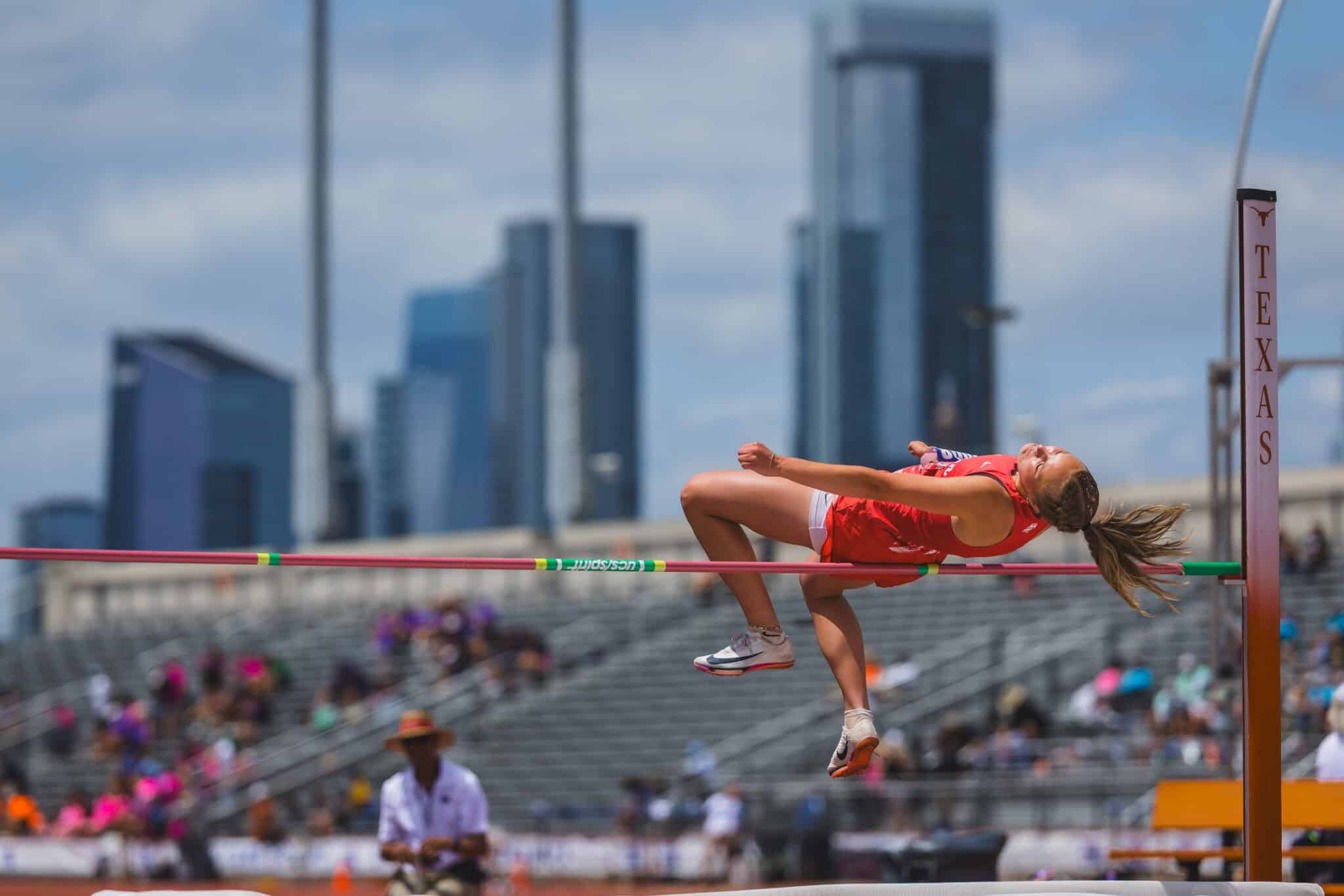 100A8689 | Press Pass Sports Female high jumper in red clears the bar during a track and field event, with a city skyline in the background.