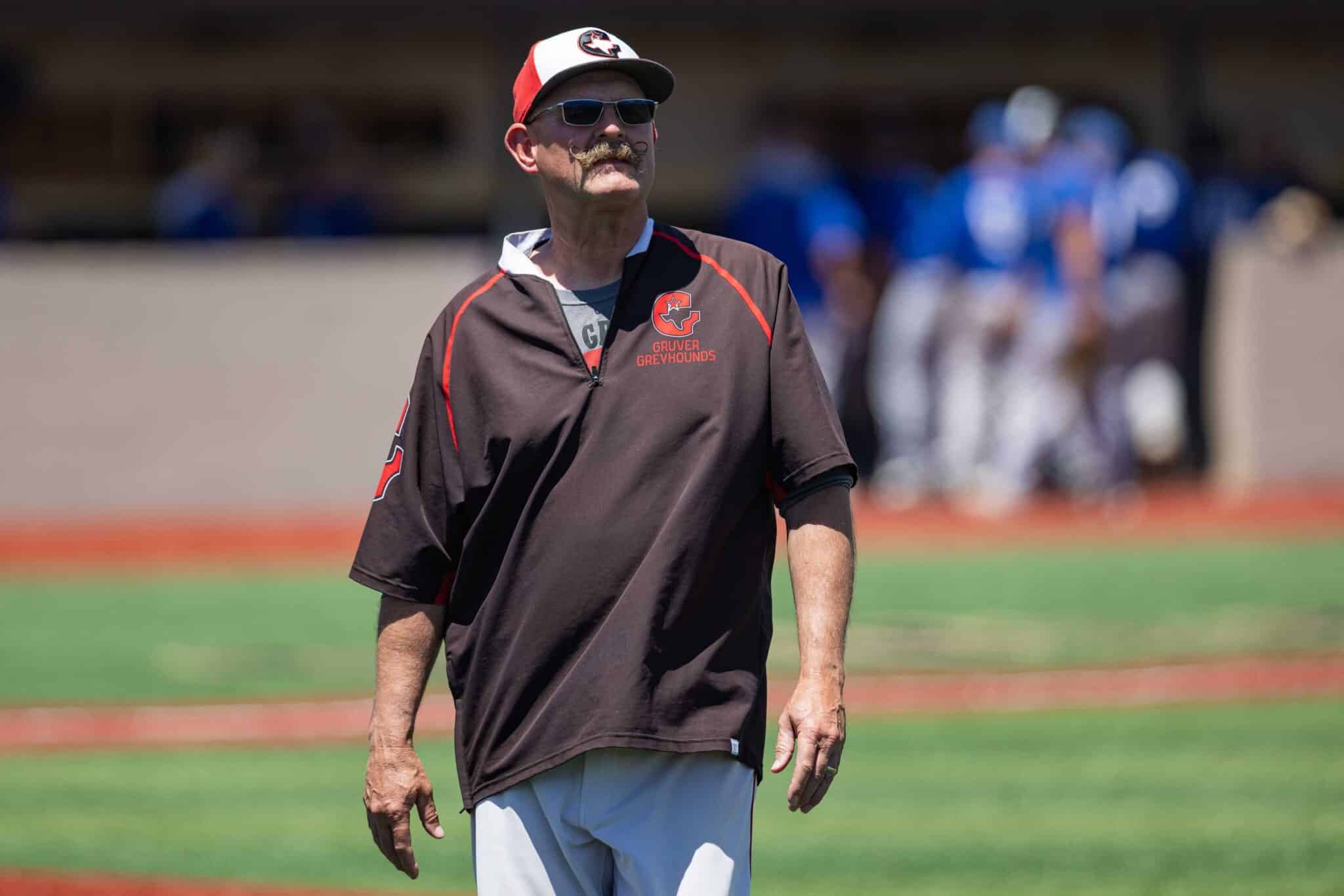 012A5278 | Press Pass Sports Mature man wearing a brown Gruver Greyhounds jacket, white cap with red visor, and sunglasses, standing on a baseball field with blurred players in the background.