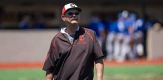 Mature man wearing a brown Gruver Greyhounds jacket, white cap with red visor, and sunglasses, standing on a baseball field with blurred players in the background.