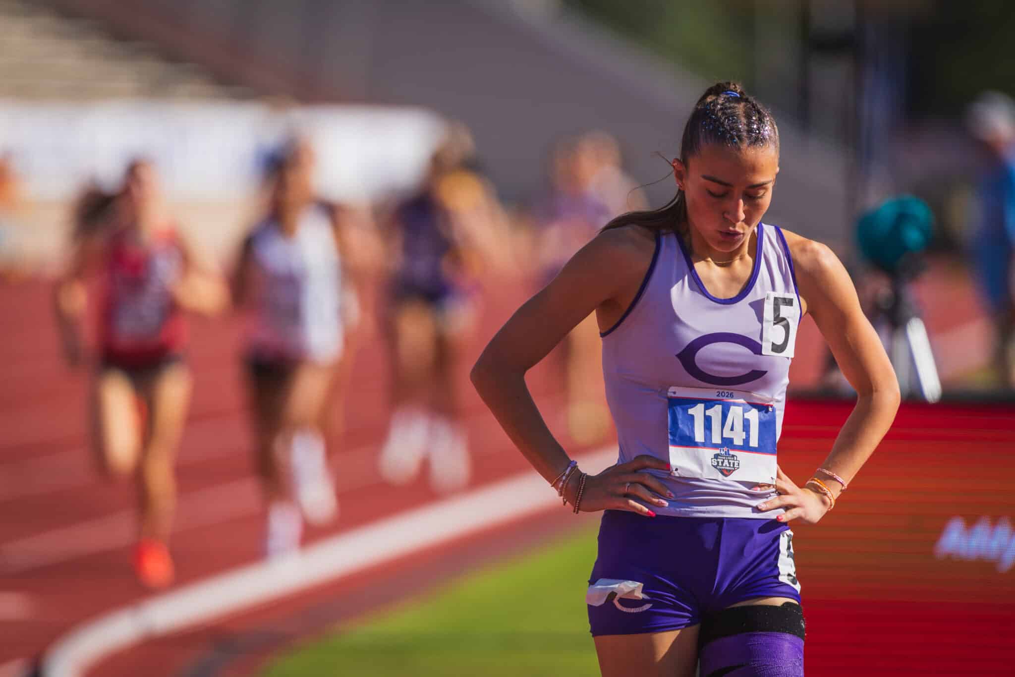 100A4747 | Press Pass Sports Female sprinter in lavender and purple with bib 1141 stands with hands on hips on a track, blurred spectators in background.