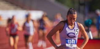 Female sprinter in lavender and purple with bib 1141 stands with hands on hips on a track, blurred spectators in background.