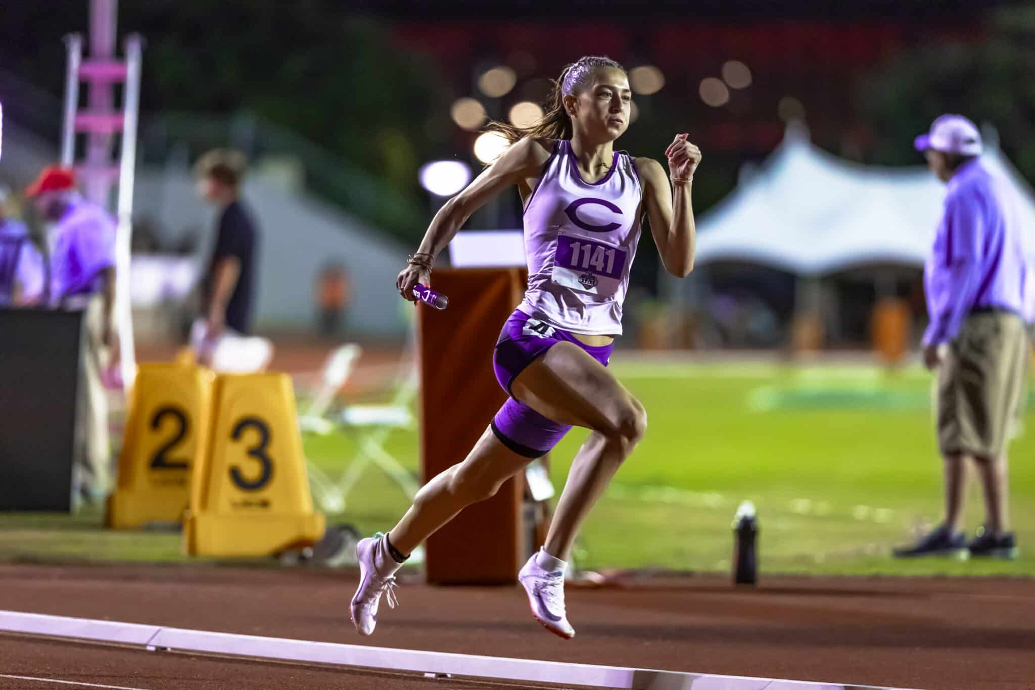 100A7584 1 | Press Pass Sports Female sprinter in purple and white kit jumping a hurdle during a night race (bib 1141).