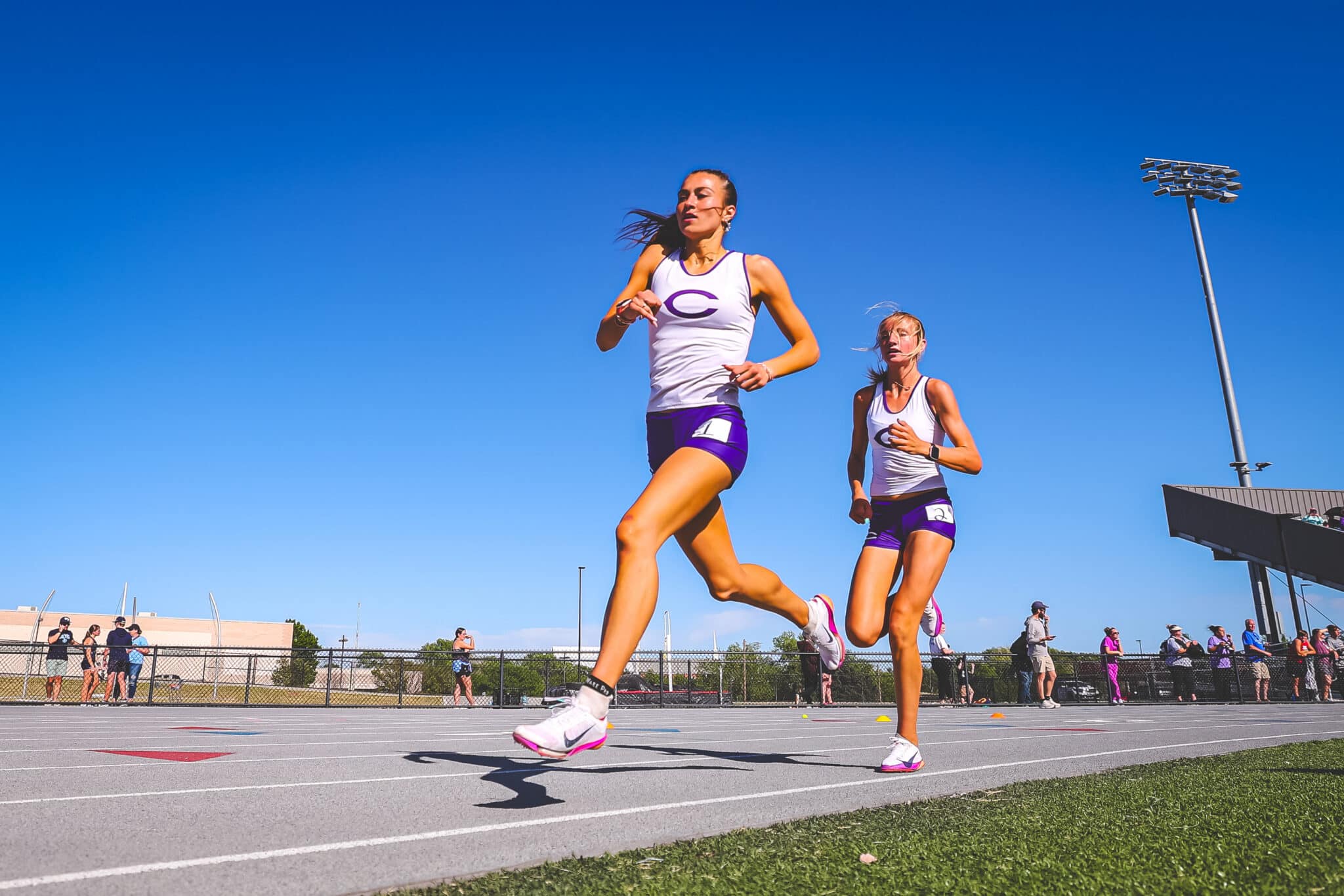 DE3_0416-2 | Press Pass Sports Two female sprinters in white and purple uniforms race down a track on a sunny day as spectators watch from the fence.