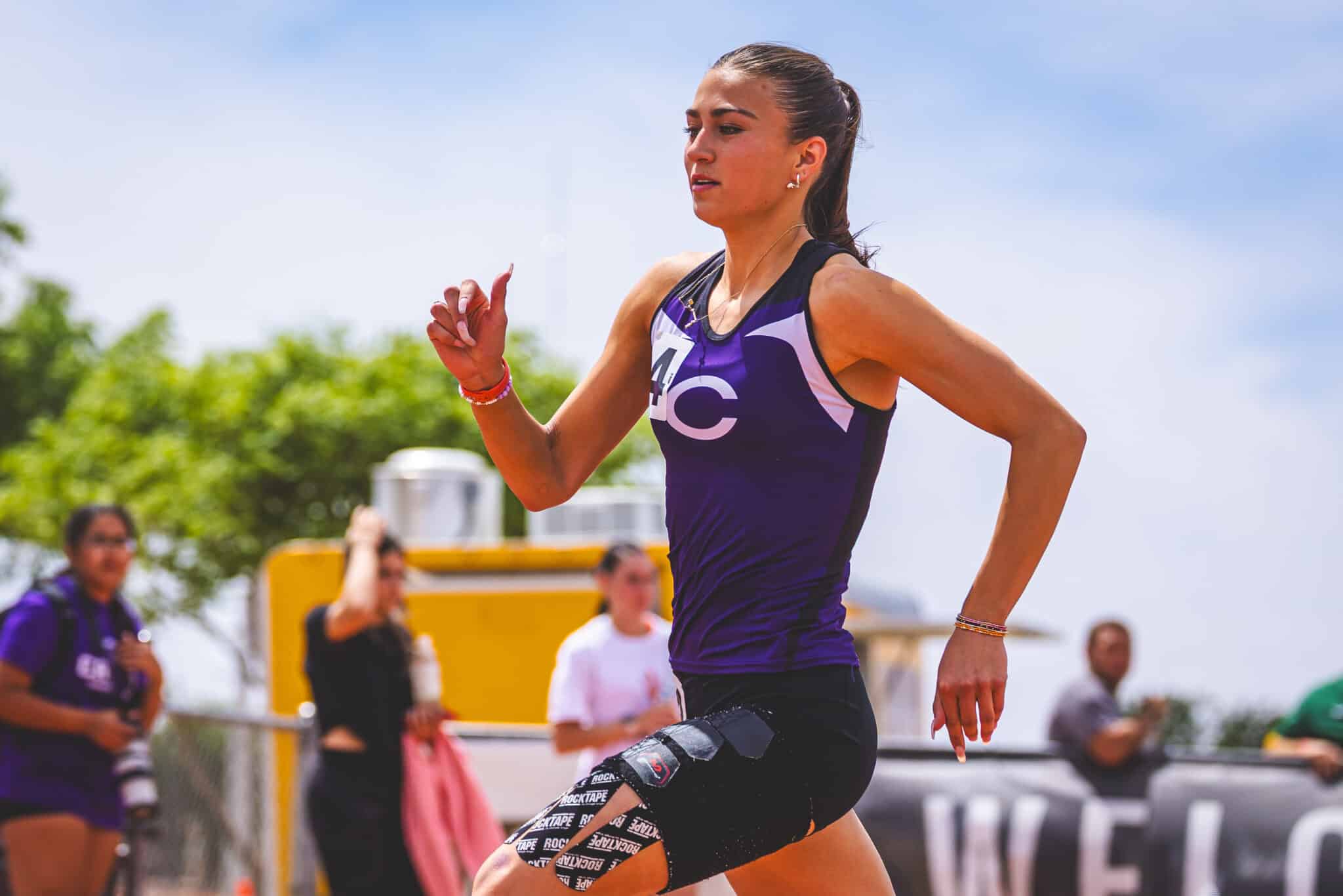 DE3_4142 | Press Pass Sports Female athlete running in a purple outfit during a race, sprinting with a focused expression and spectators in the background.