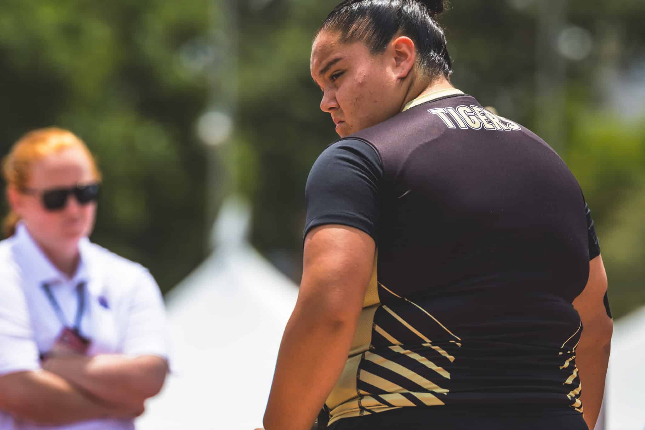 Athlete wearing a black and gold uniform with 'TIGERS' on the back, facing away outdoors; a blurred person in sunglasses in the background.