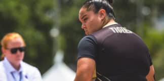 Athlete wearing a black and gold uniform with 'TIGERS' on the back, facing away outdoors; a blurred person in sunglasses in the background.