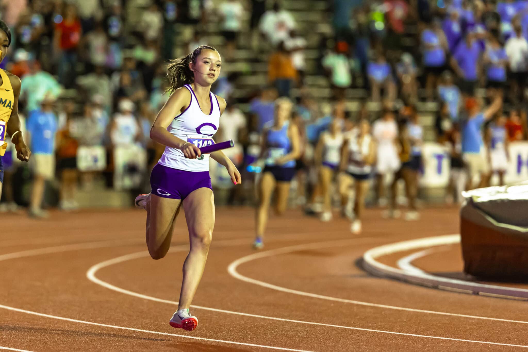 Female athlete running with a baton on a track during a relay, with crowded stands in the background.