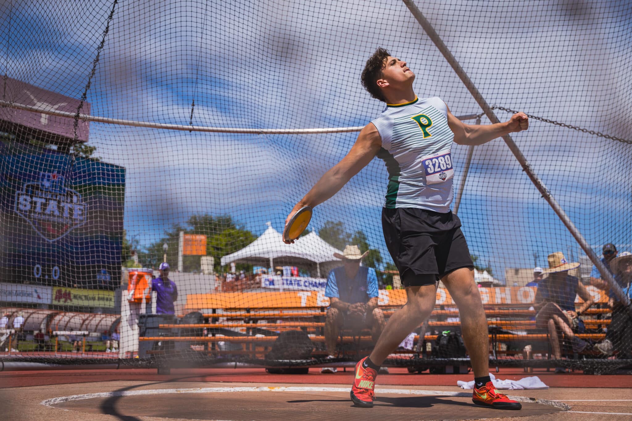 Male athlete in a white-green jersey with bib 3289, sprinting inside a fenced court during a race.