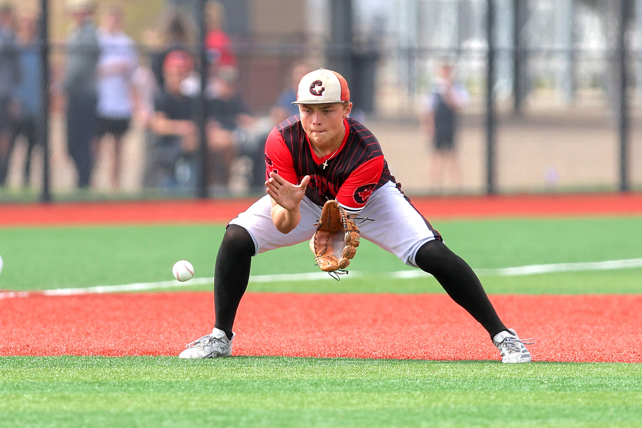 Baseball pitcher in red and black uniform extends to field a ground ball, glove open as the ball rolls toward him on a bright field.