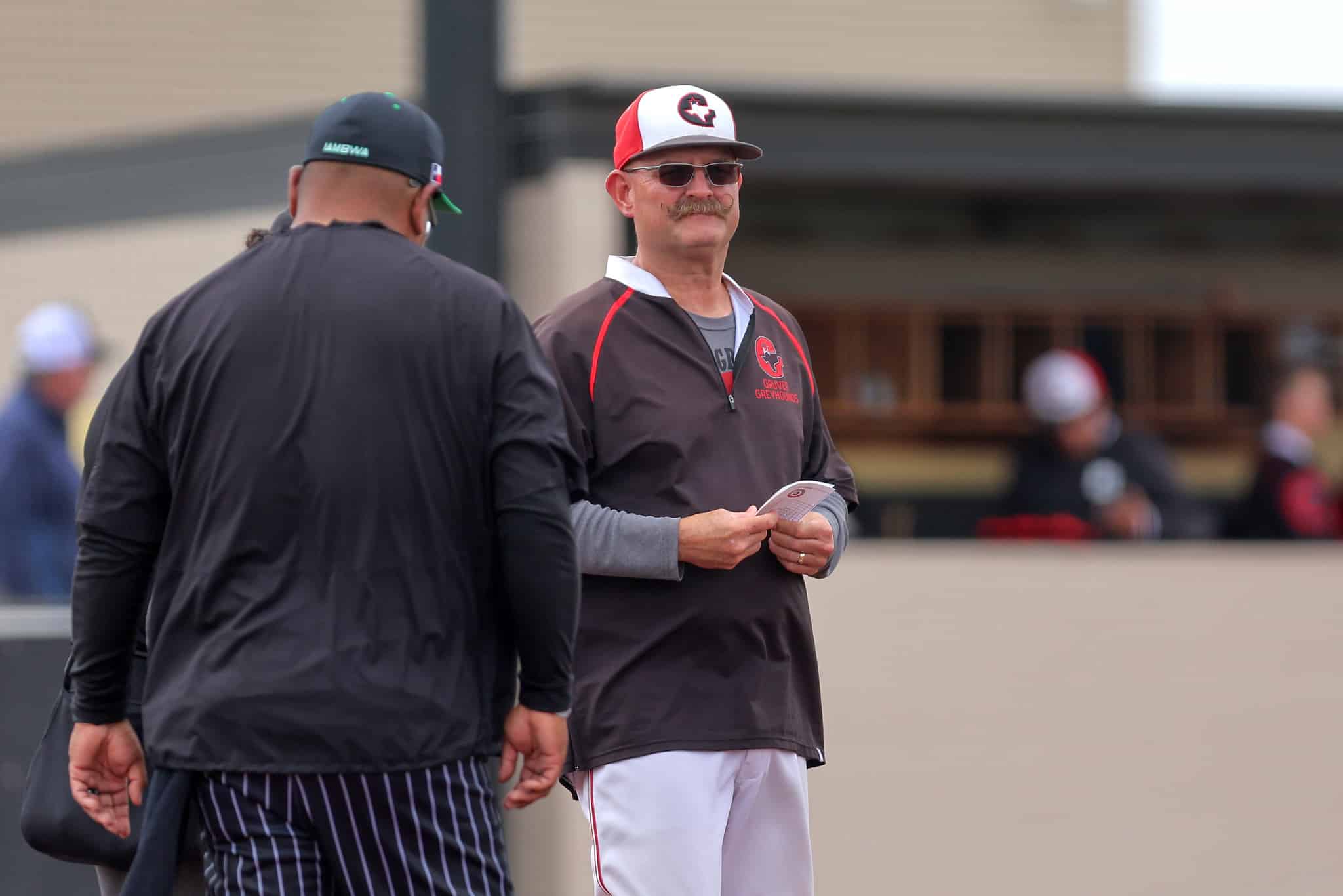Two men talking outdoors; one faces the camera in a gray jacket and red-and-white cap, the other with his back turned in a dark jacket.