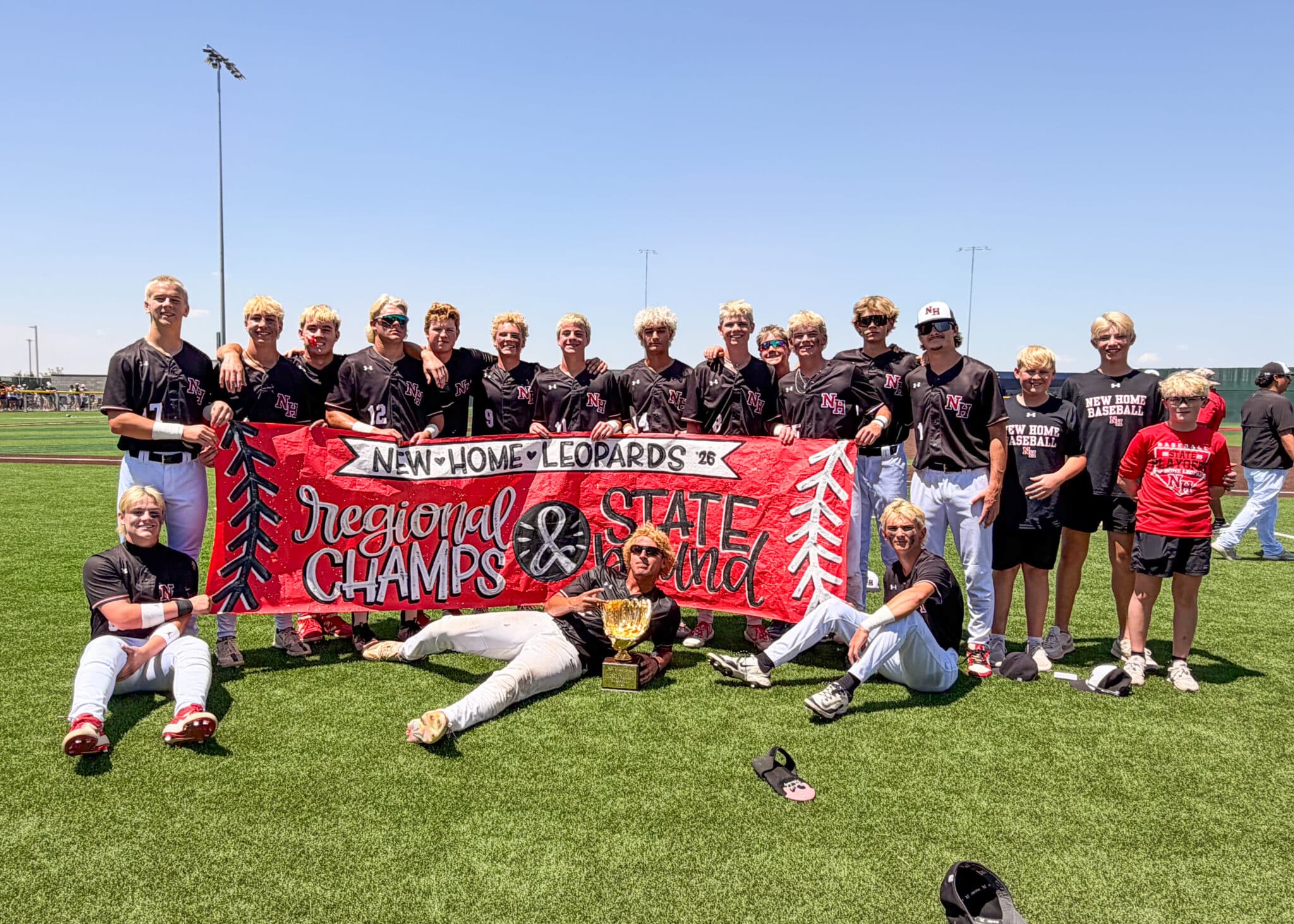 IMG_5197 1 | Press Pass Sports Group of young baseball players in black uniforms pose behind a red banner on a sunny field, trophy in front, celebrating a win.