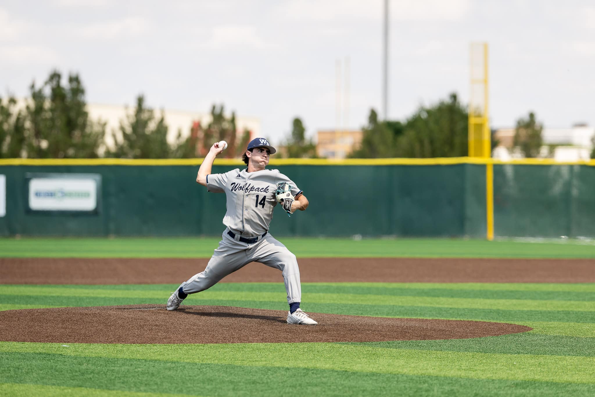 west-plains-baseball-jpegJoe-pps-3 | Press Pass Sports Baseball pitcher in gray Wolfpack uniform throwing a pitch from the mound on a sunny field.