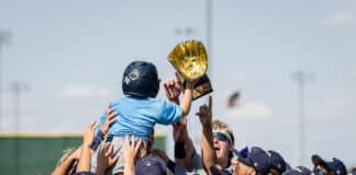 Baseball players in gray uniforms celebrate, lifting a young child on shoulders while holding a large gold trophy overhead.