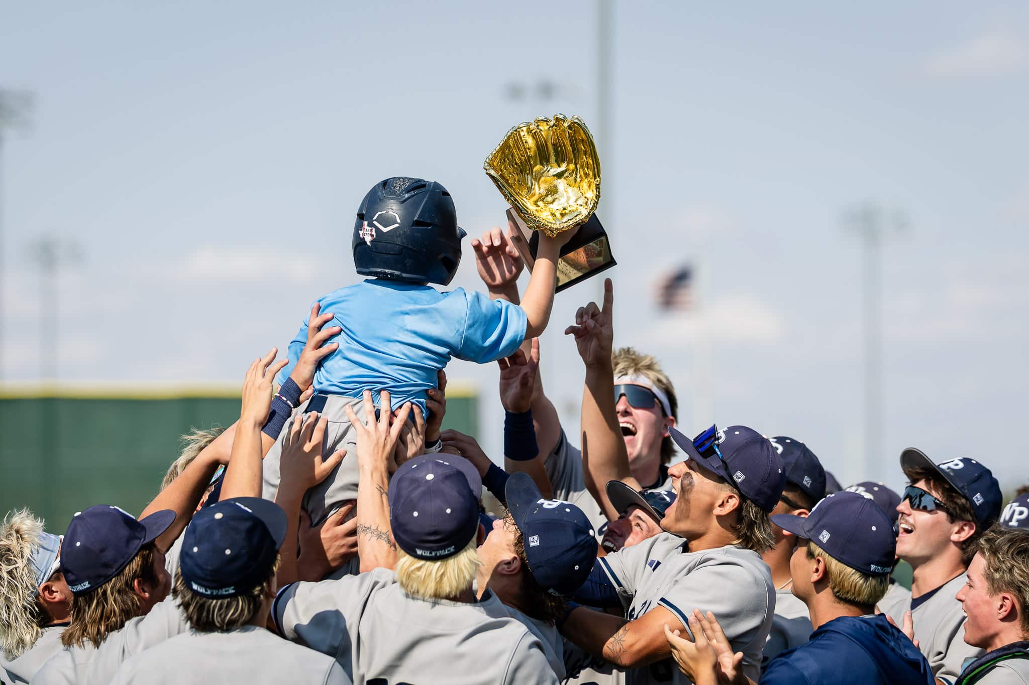 west-plains-baseball-jpegJoe-pps-11 | Press Pass Sports Baseball players in gray uniforms celebrate, lifting a young child on shoulders while holding a large gold trophy overhead.