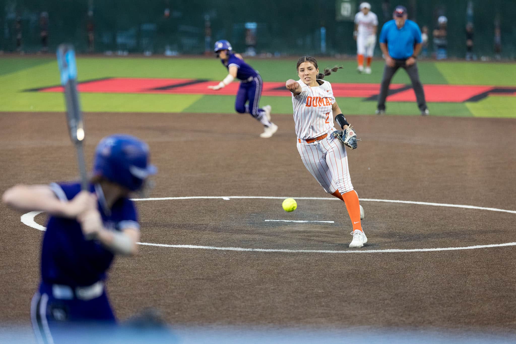 dumas-softball-jpegjoe-pps-7 | Press Pass Sports Female softball pitcher in white pinstripes delivers a pitch toward home plate, with a blue-uniform batter in foreground and players in the background.