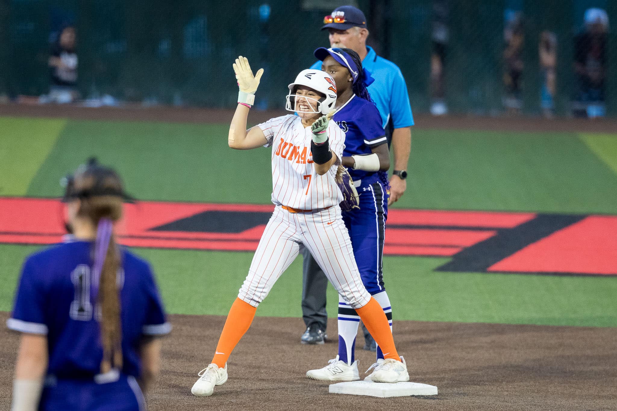 dumas-softball-jpegjoe-pps-10 | Press Pass Sports Softball player in white pinstriped uniform with orange socks on first base, raising arms in celebration as teammates and an umpire watch in the background